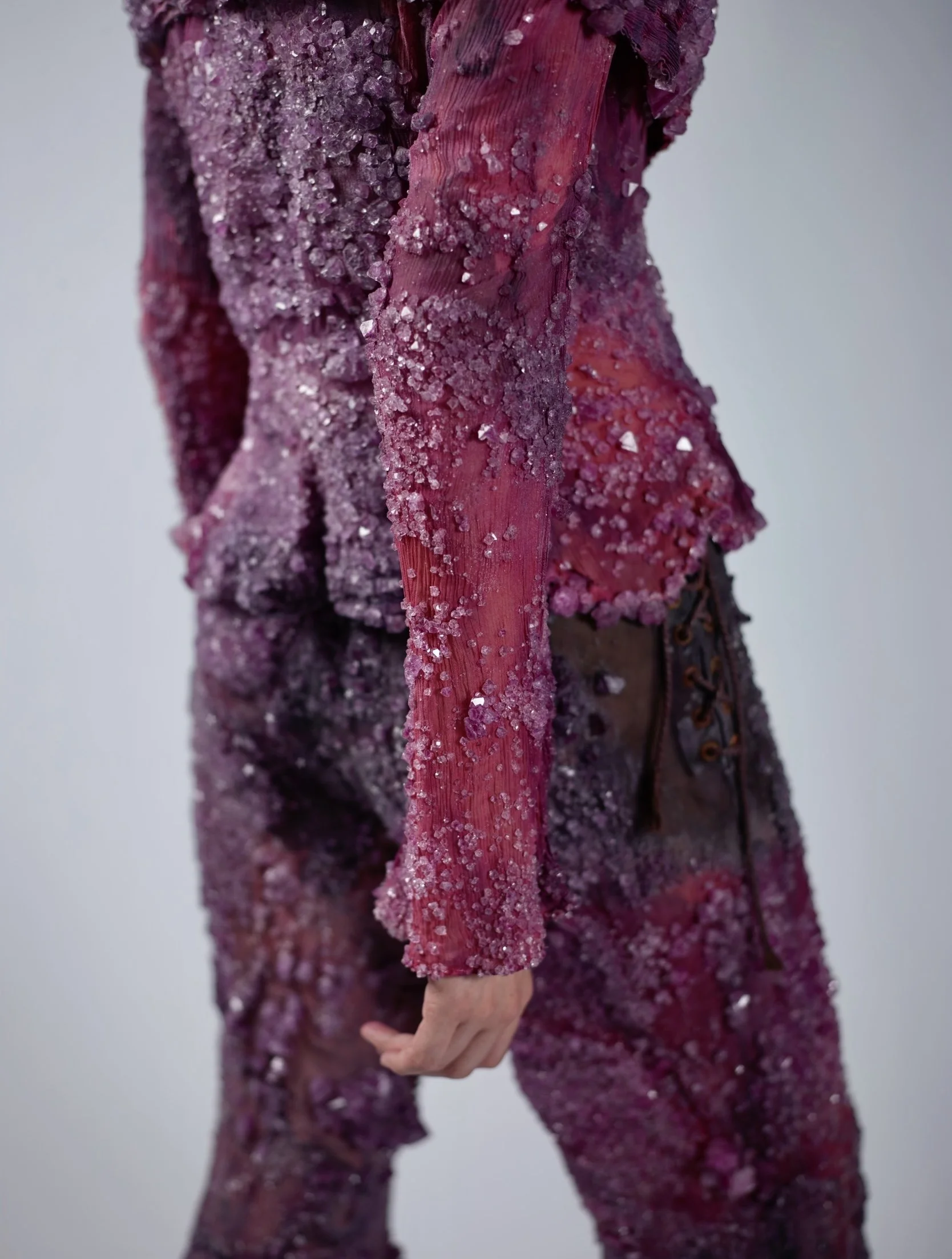 Close-up of a person wearing a purple, glittery, textured outfit with salt crystals on the fabric, holding their hand behind their back.