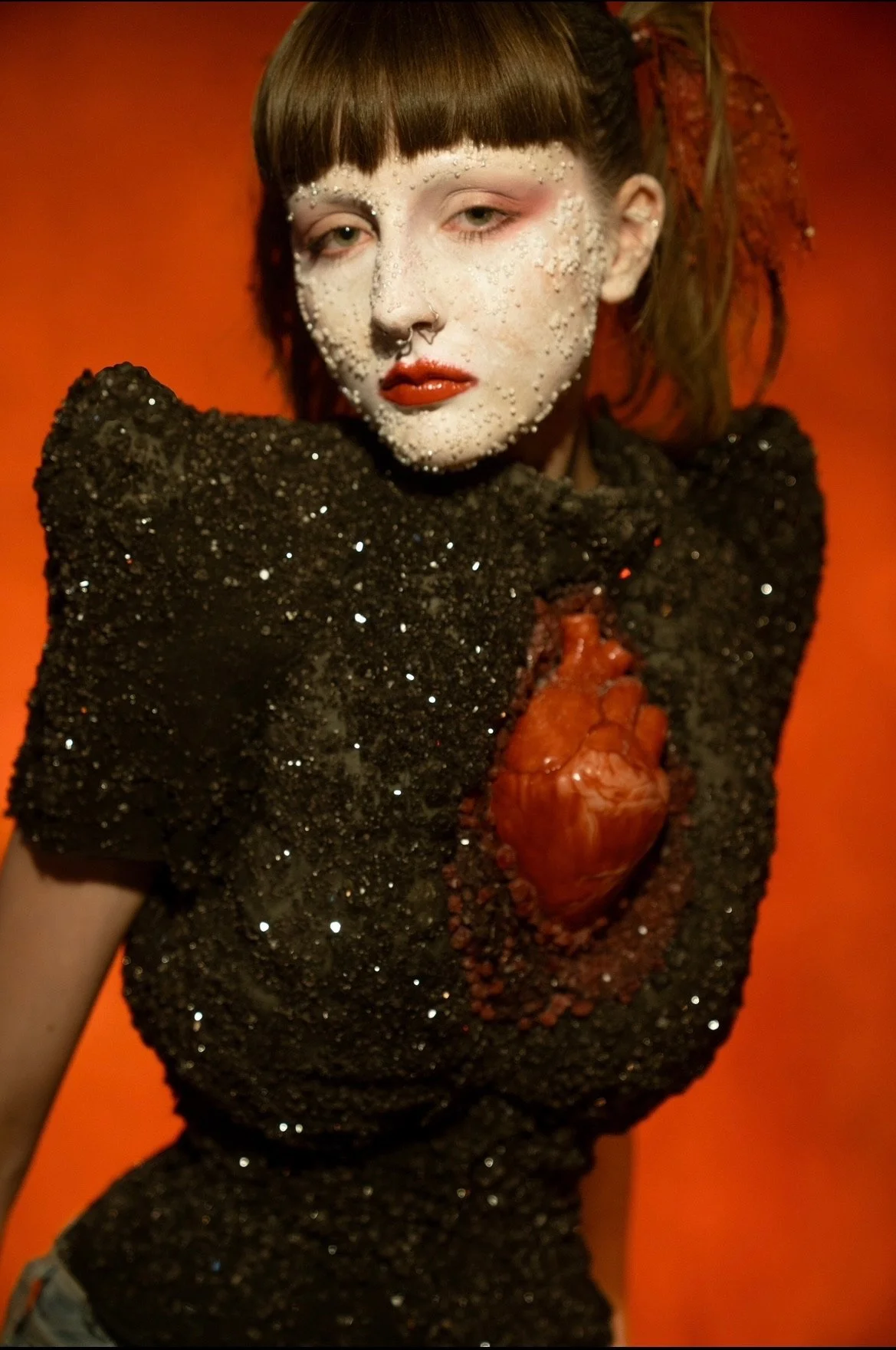 A woman with pale makeup, bubble-like decorations on her face, and dark lipstick. She has brown hair styled with braids and wears a black sequined dress with a design resembling a human heart in the chest area. She poses against an orange background.