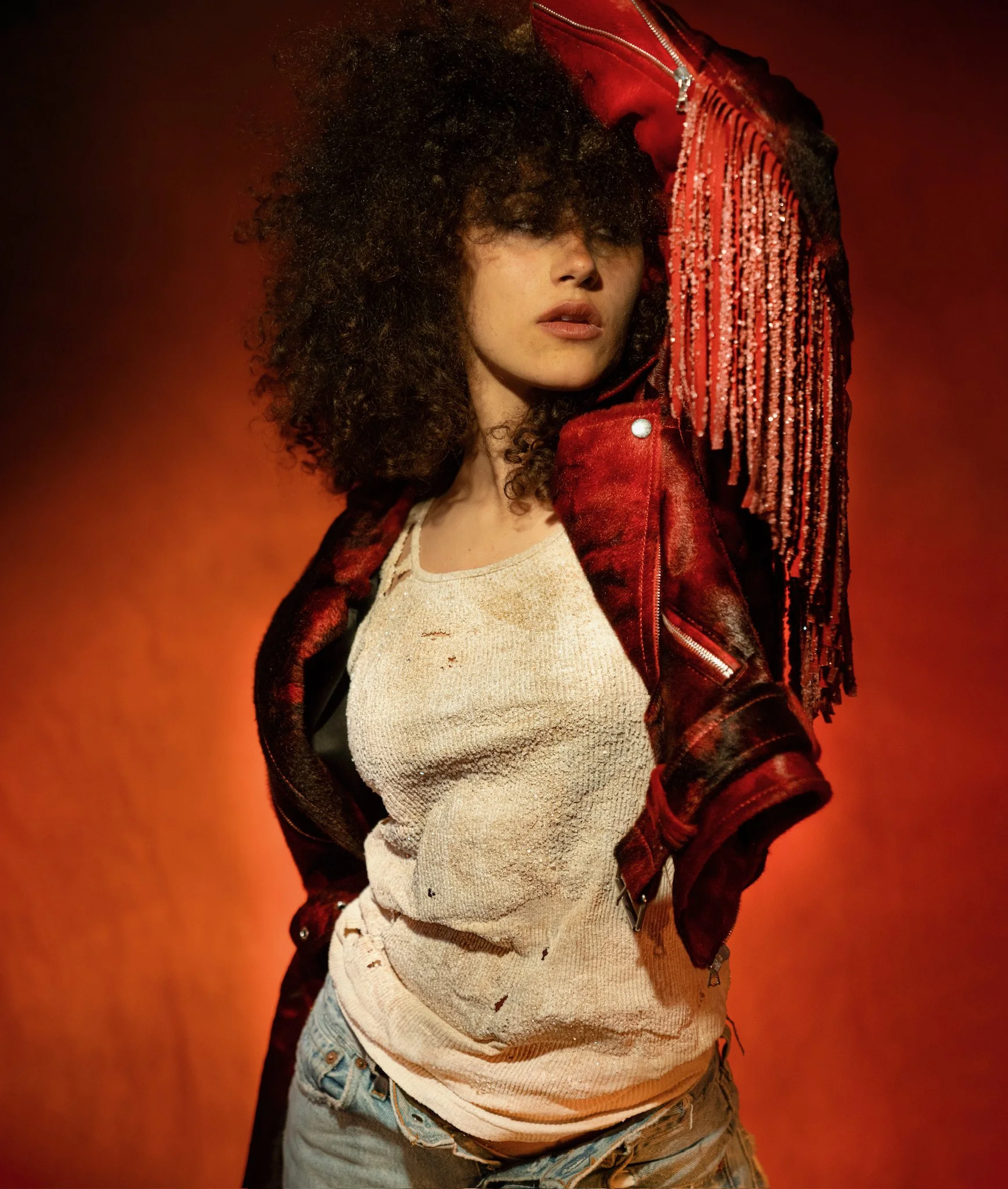 A woman with curly hair wearing a distressed beige tank top, a red leather jacket with fringe details, and ripped jeans stands against a reddish-orange background.