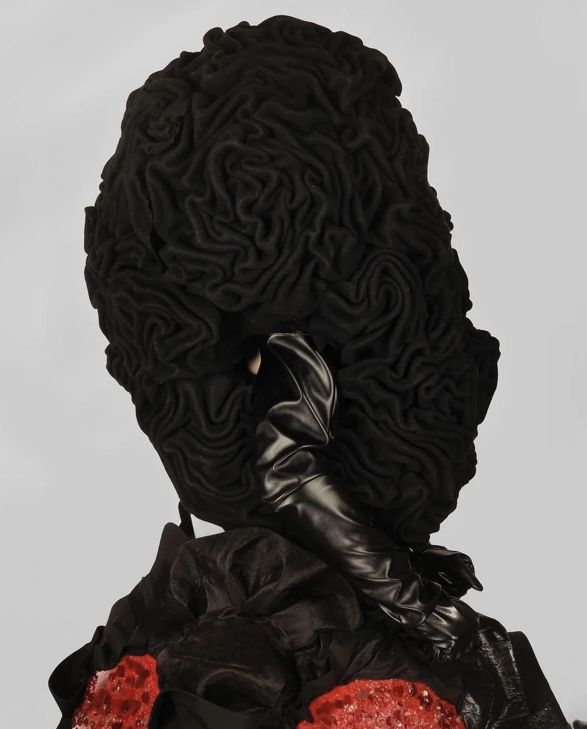 An artistic representation of a human head made from moroccan fabric resembling brain folds, wearing a black leather glove covering the mouth, with a black fabric and red lace necklace or fabric at the base.