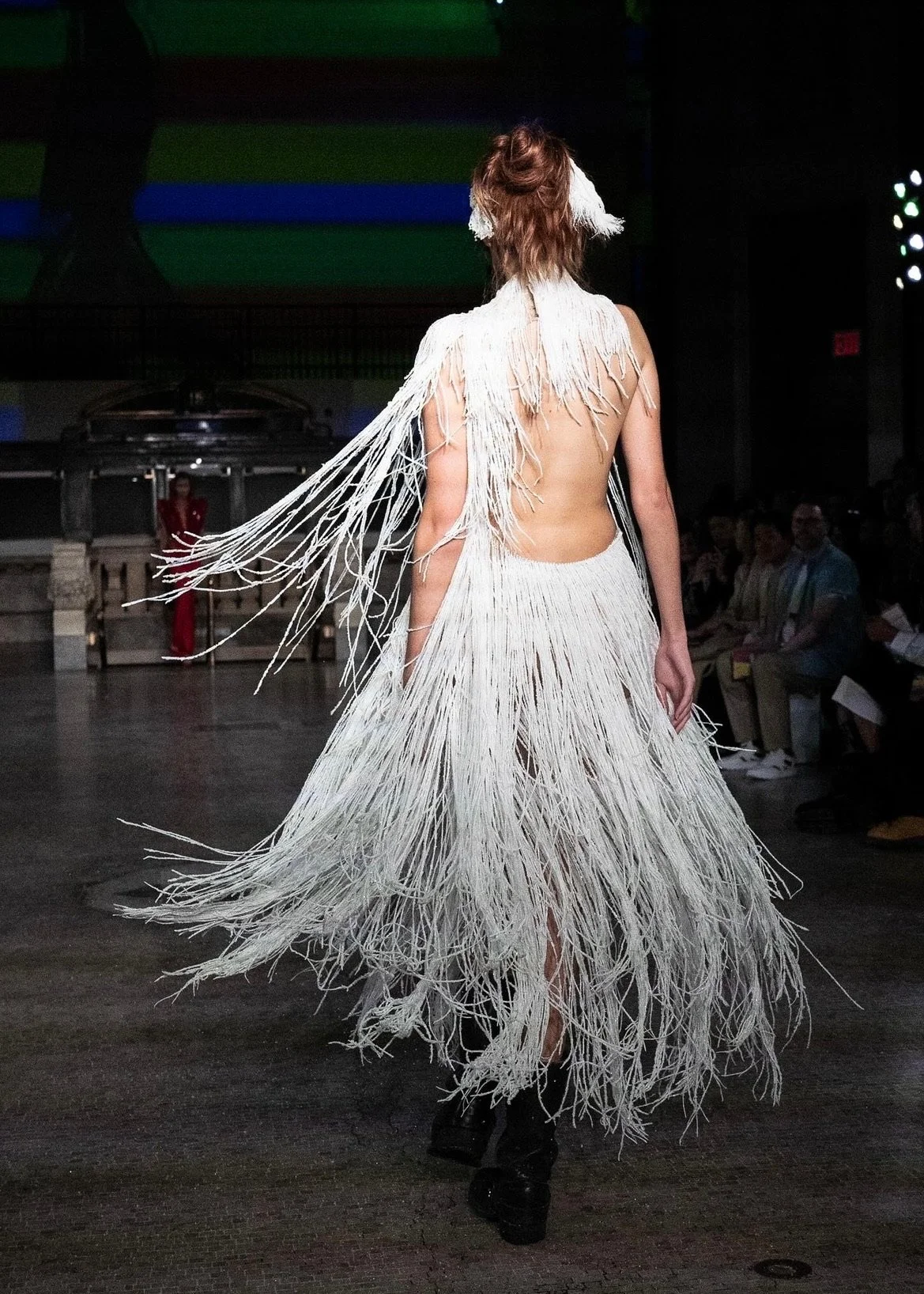 A model walking on a runway wearing a white, fringe-covered dress with a backless design and matching accessories in a fashion show.