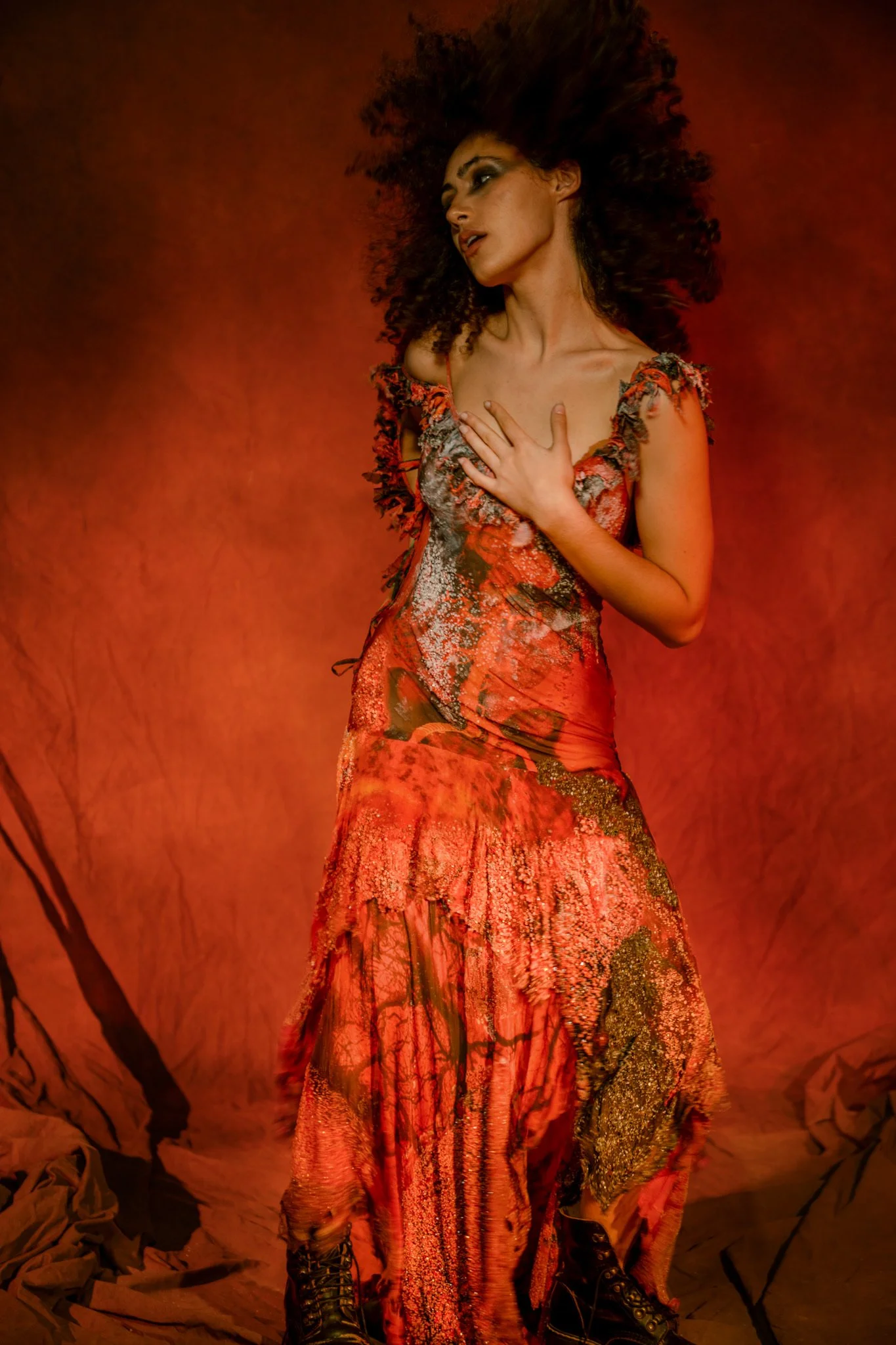 A woman with curly hair wearing a flowing, multicolored dress with a floral pattern, standing against an orange backdrop.