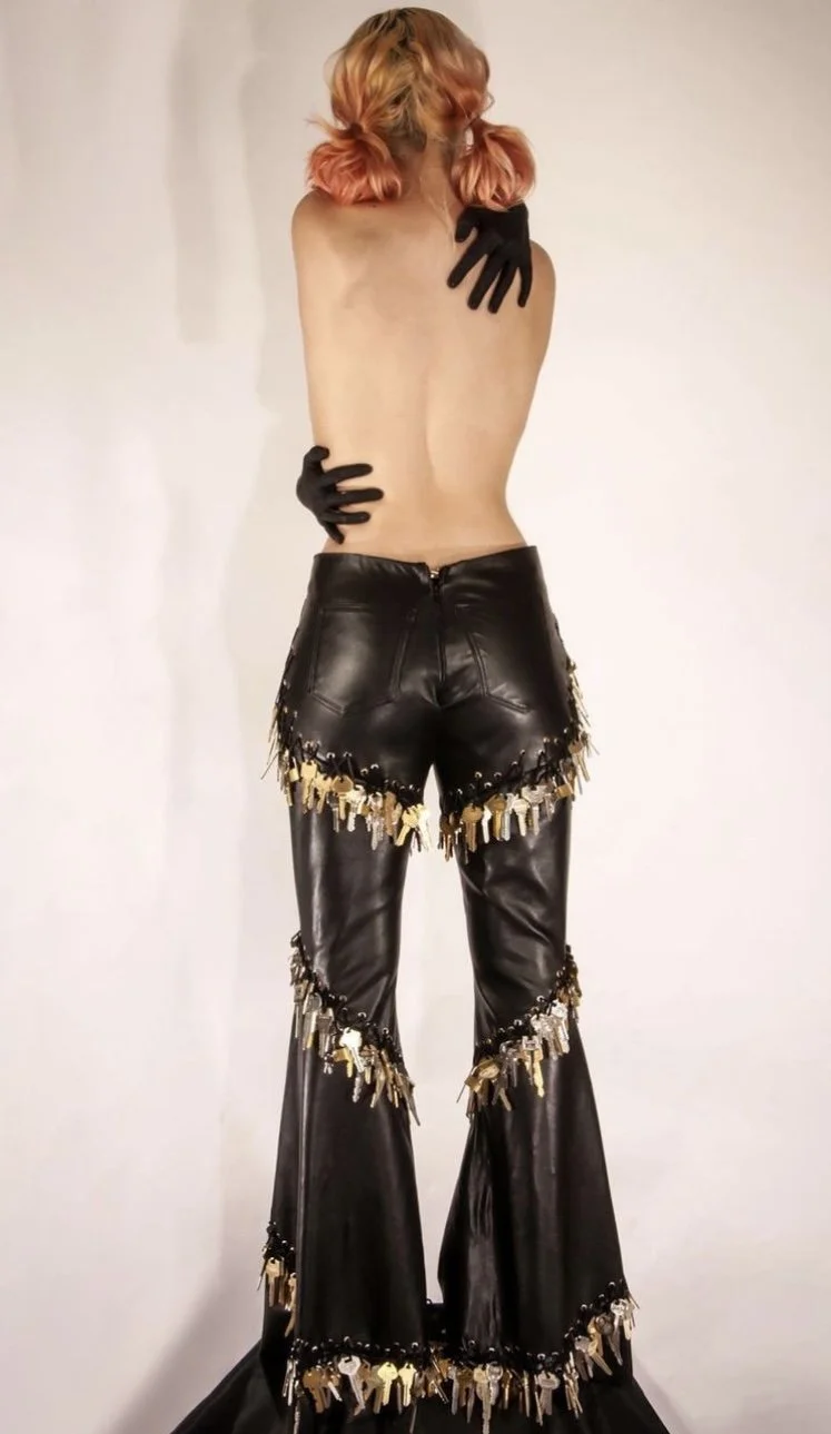 Person with pink hair, topless, wearing black gloves, black leather pants with gold hardware, standing against a plain white background.