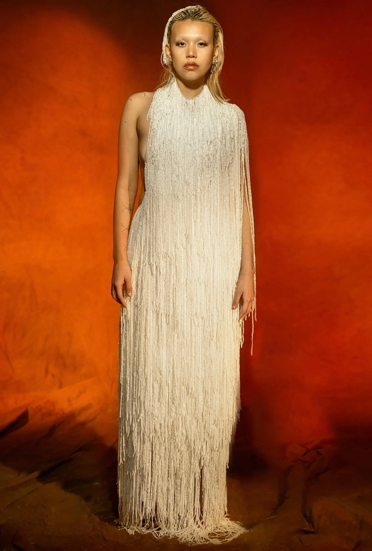 A woman with blonde hair and light makeup wearing a long, sleeveless, cream-colored dress with fringes, standing against an orange background.