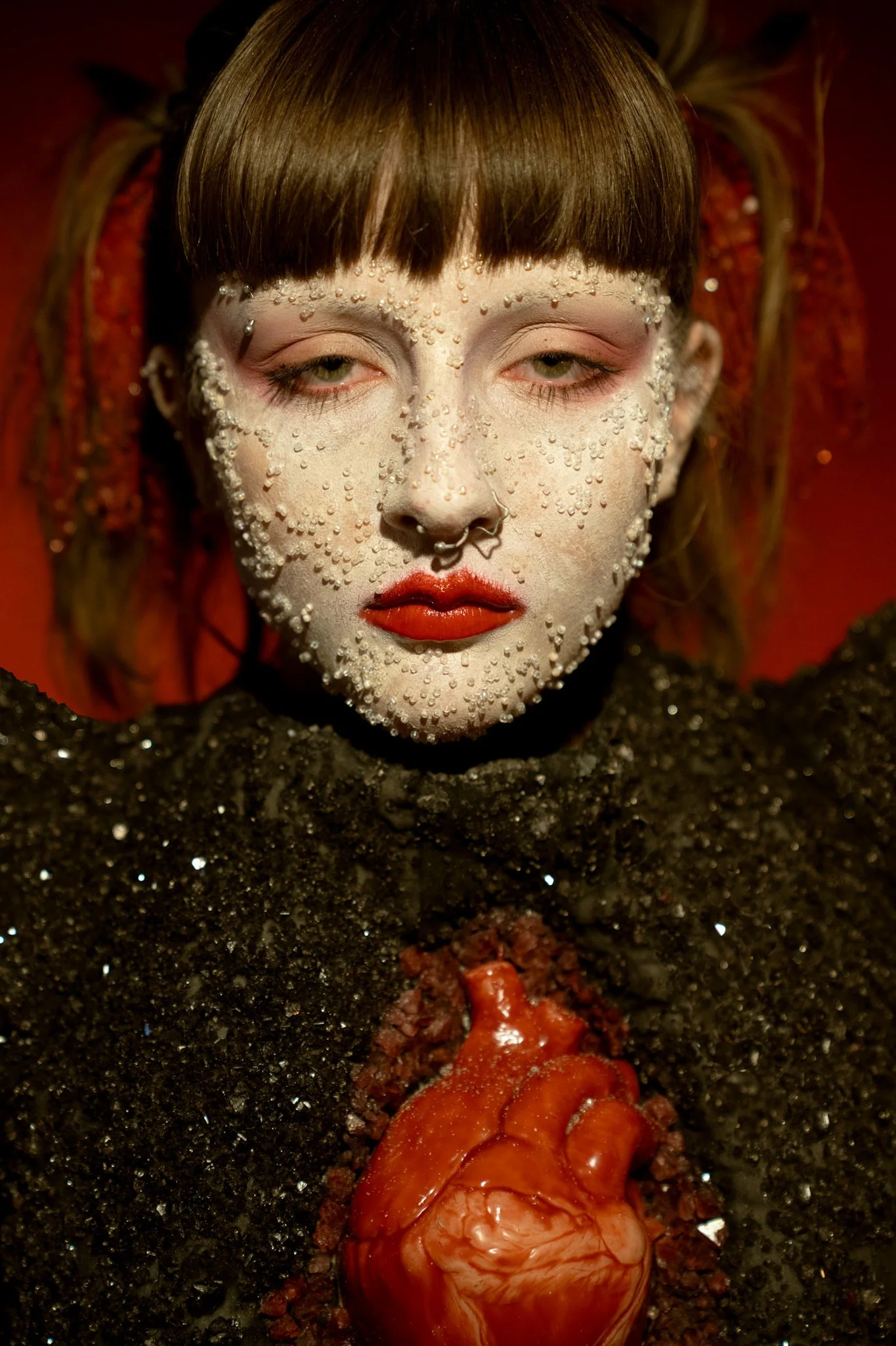 A person with a pale face decorated with small white beads and red lipstick, wearing dark clothing and a black choker. They have a septum piercing shaped like a star and are standing in front of a red background. A heart is placed on their chest, whi