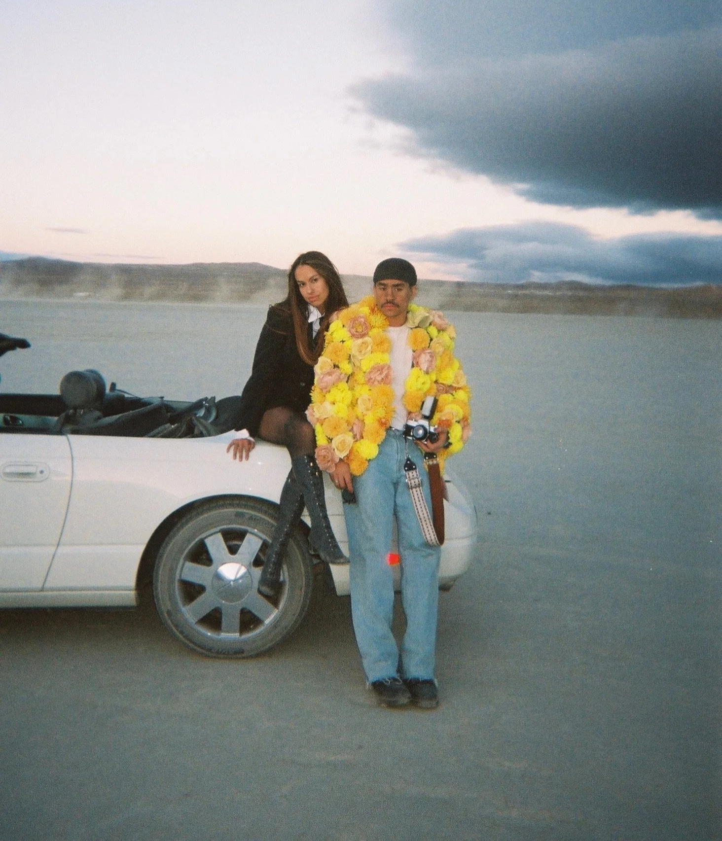 Young man in light blue jeans and floral jacket standing beside a white convertible car, with woman in dark clothing sitting on the car's door, near a large body of water under cloudy sky.