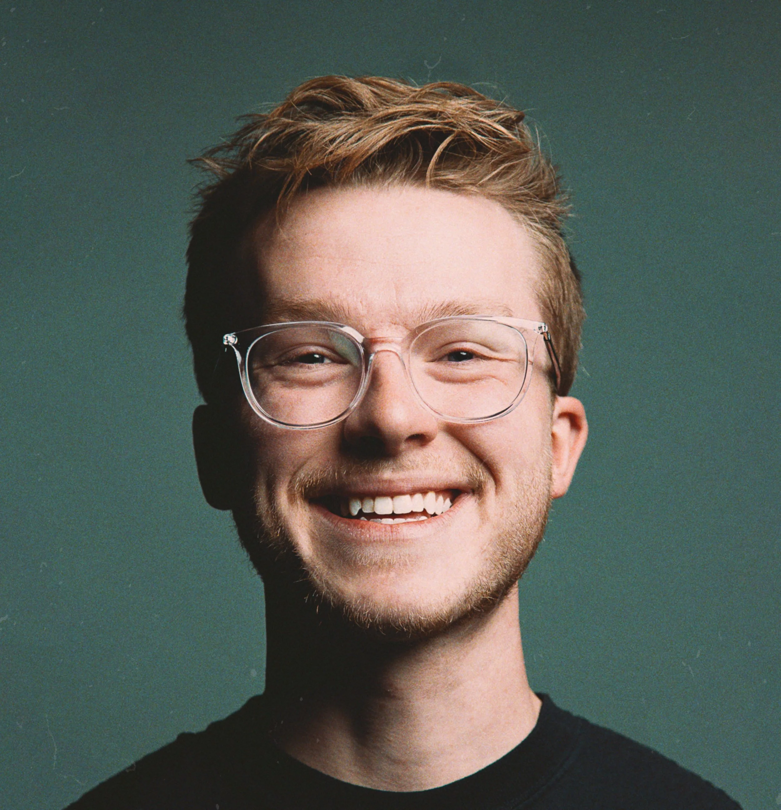 Close-up of a young person's face showing a cheerful smile, glasses, short brown hair, and light skin, against a dark green background.
