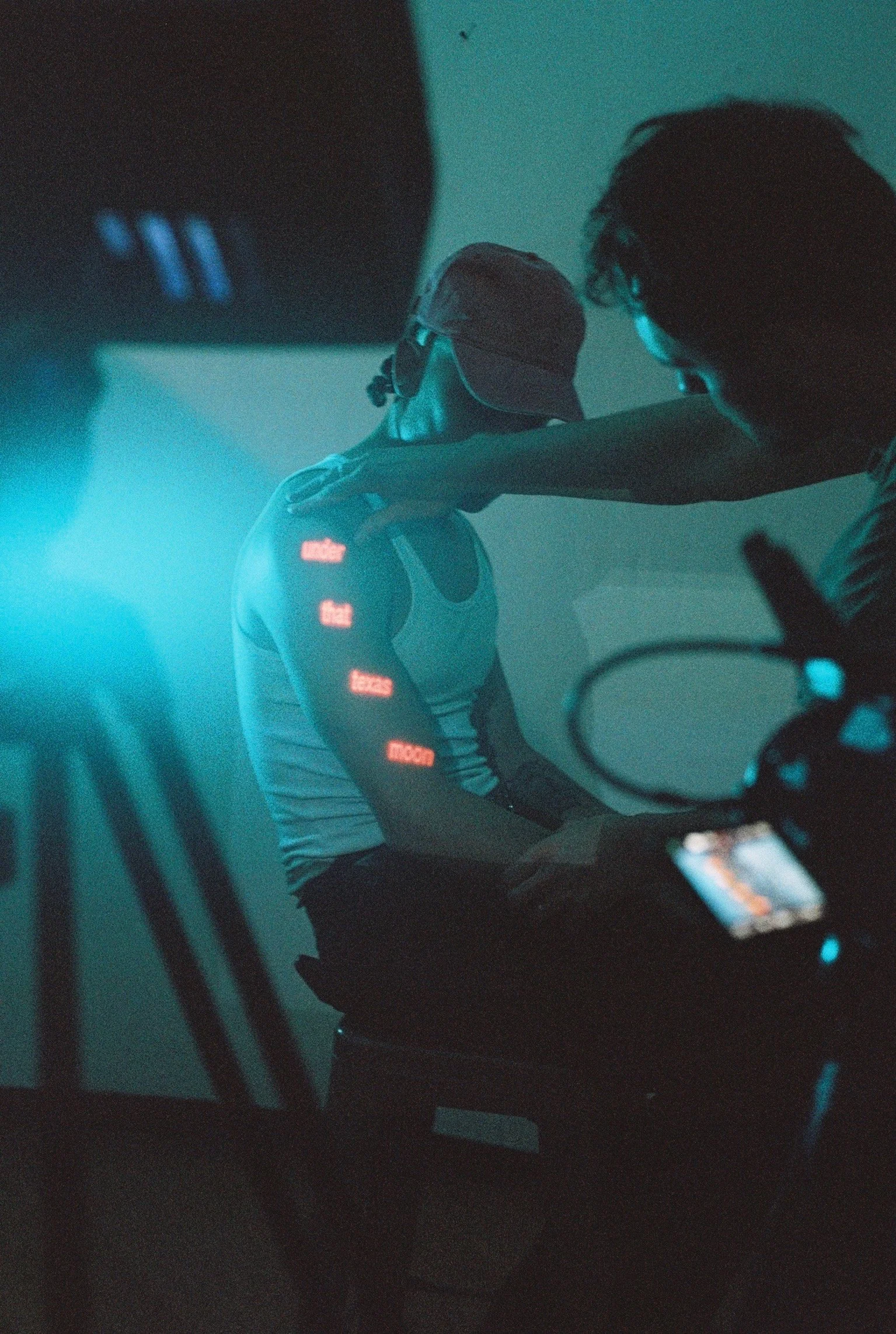 A person wearing a baseball cap and headphones, sitting with their head resting on their arm, which is placed on a table. The scene is dimly lit with a blue-green color tone.