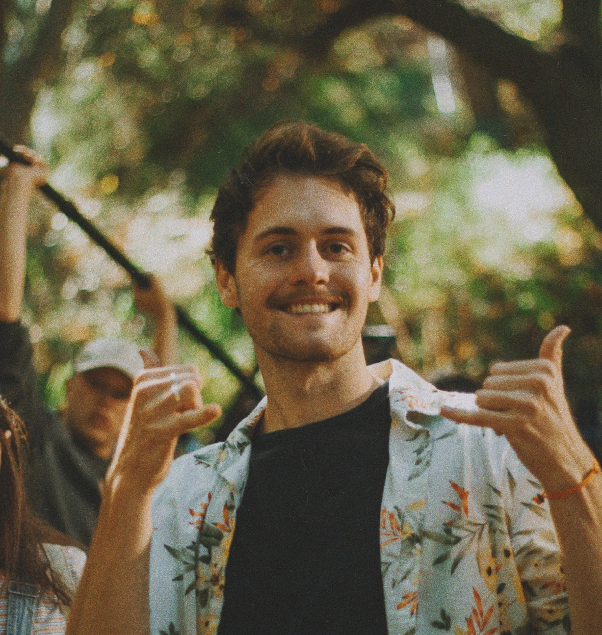 A young man with brown hair and a beard smiling and making a hand gesture, outdoors with blurred trees in the background.