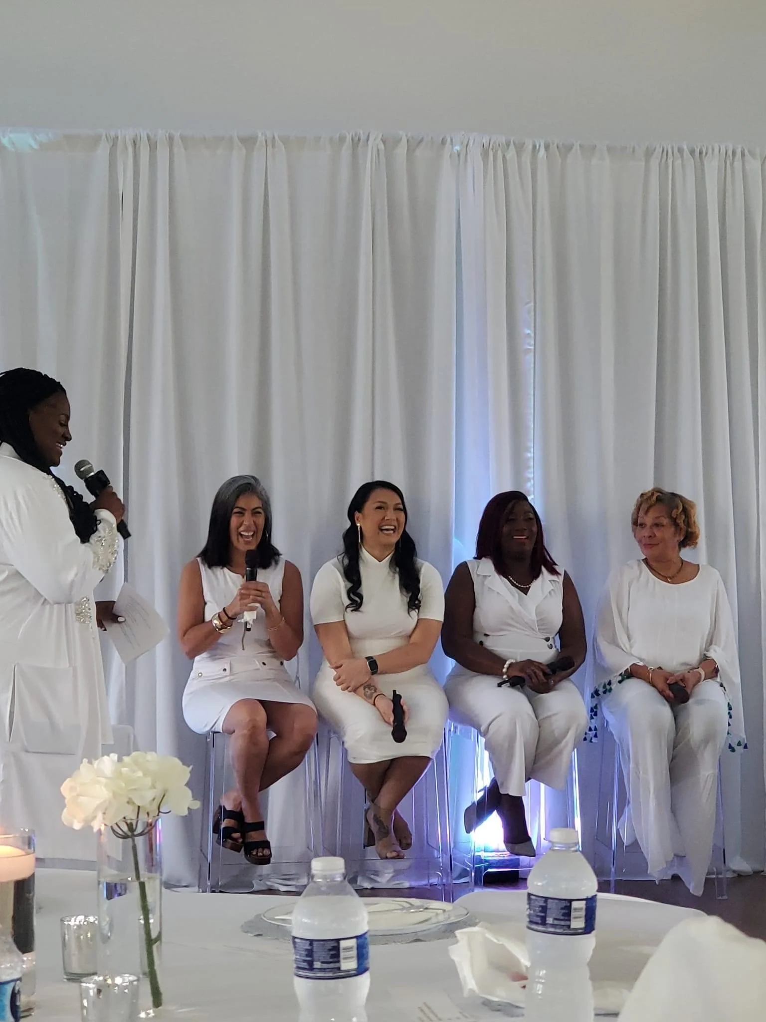Five women dressed in white sitting on high chairs on stage during an event, with a woman standing and speaking into a microphone. The background has white curtains, and there are water bottles and floral arrangements on the table in front.