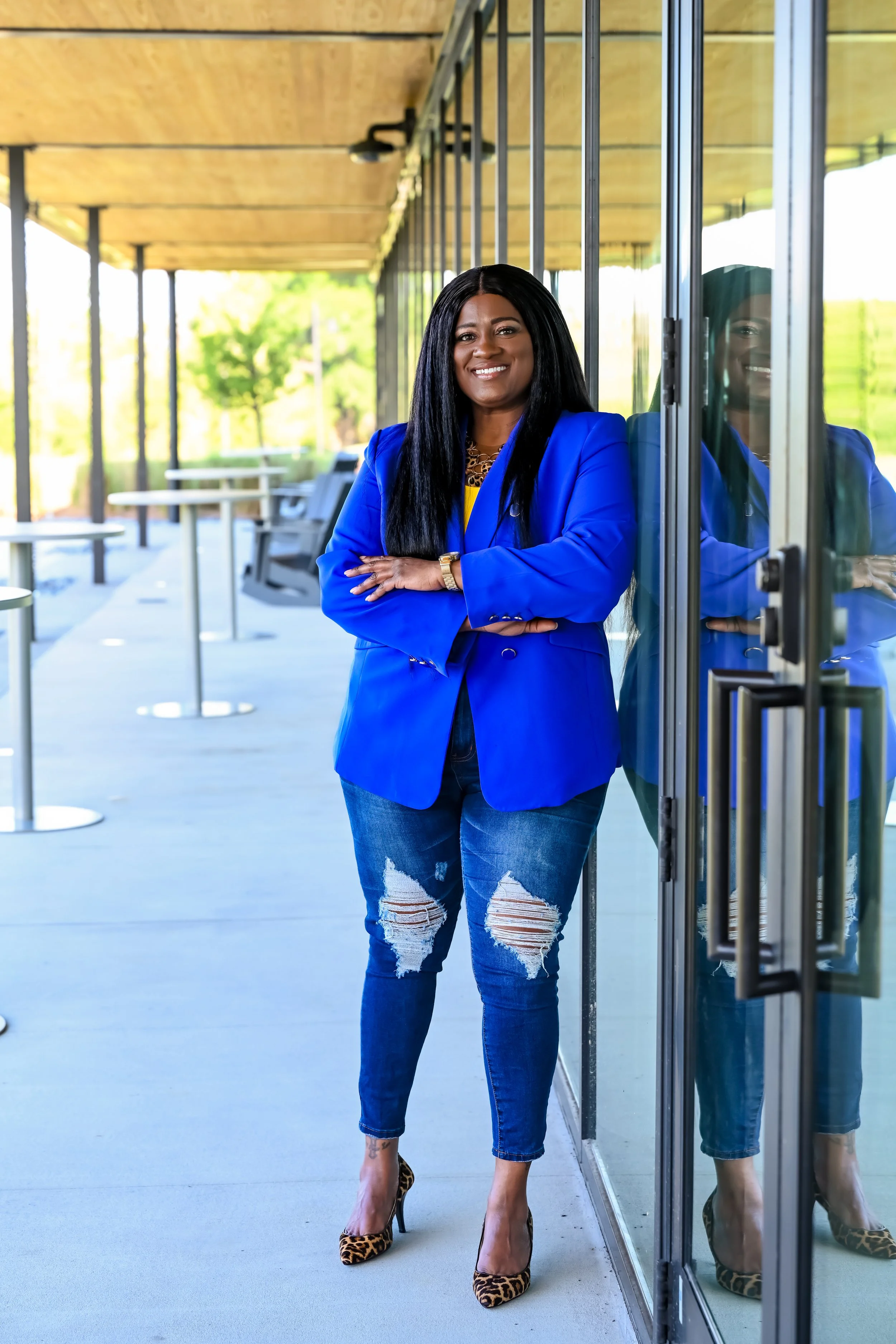 A woman in a bright blue blazer and ripped jeans standing outside a building with glass walls, smiling with cross arms.