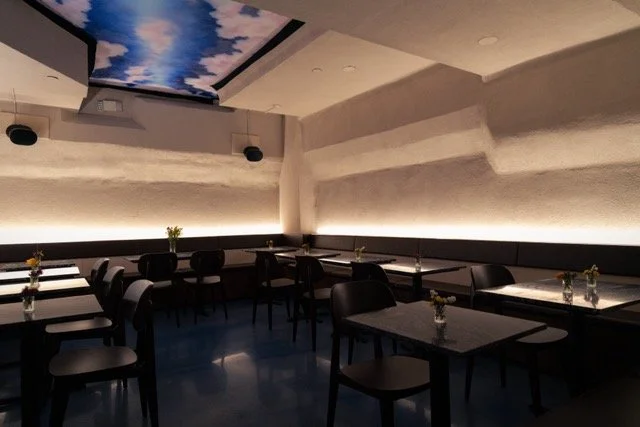 Empty restaurant with black tables and chairs, small vases with flowers, dim lighting, and a ceiling painting of a cloudy sky.