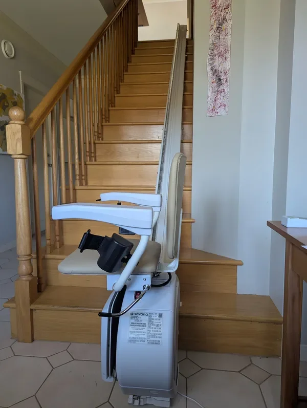 Stair chair lift installed on wooden staircase in a home, with beige upholstered seats and wooden handrails.