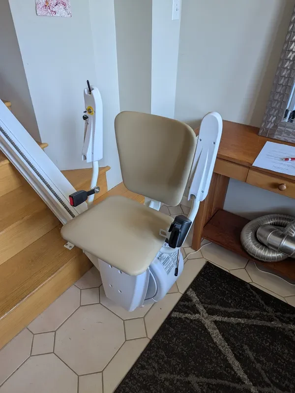 A beige stairlift chair with a cushioned seat and backrest in a corner of a room, next to a staircase and a wooden desk with papers and a window.