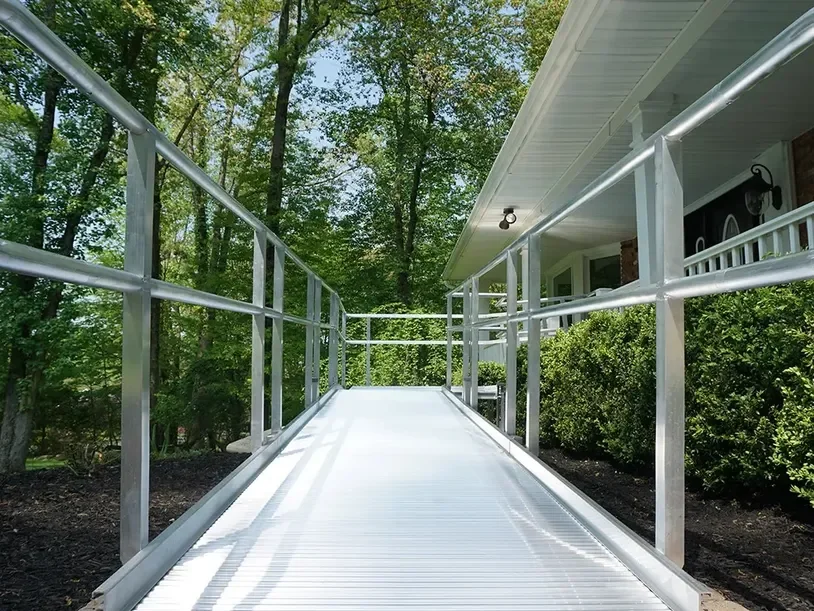 View of a metal wheelchair ramp with safety rails leading to the house porch, surrounded by trees and bushes.