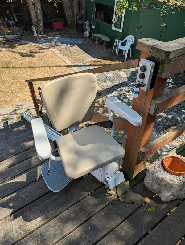 An outdoor stairlift chair mounted on wooden steps outside a house, with control switches on the side and a cushioned seat.
