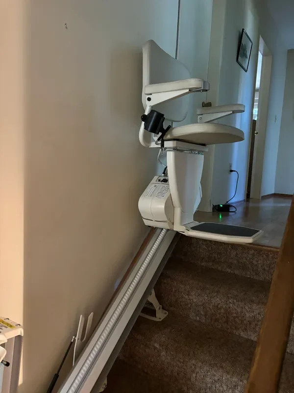 A stair lift with a beige seat and backrest installed along a staircase, positioned next to a beige wall in a home interior.