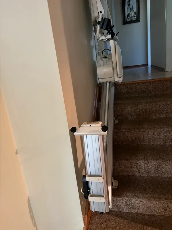 A stairlift chair mounted on a staircase with brown carpet. The stairlift has a white seat and black safety arms, positioned at the bottom of the stairs.