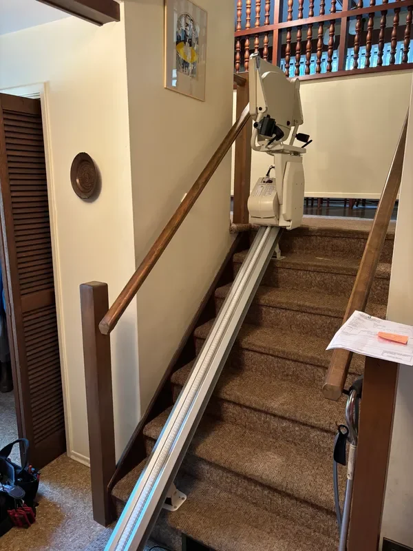 A stairlift installed on a staircase, with a chair at the upper landing and a rail running down the steps, in a home interior.