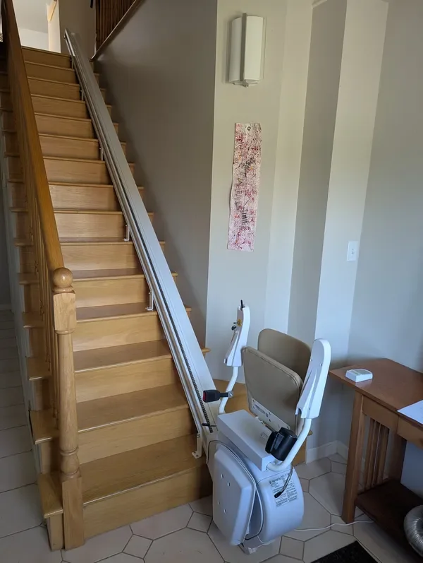 A stair lift installed on a wooden staircase in a home. The stair lift has a beige seat and metal rails running along the side of the stairs. There is a white control box and a footrest at the bottom.