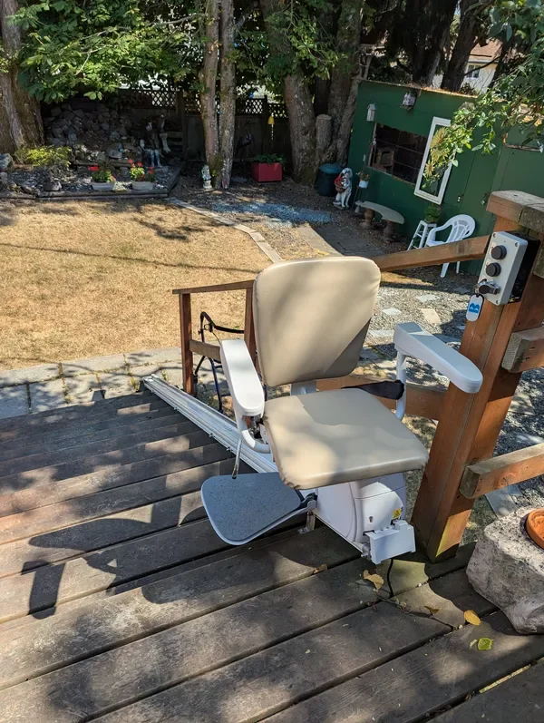 A stairlift chair on a wooden deck overlooking a yard with trees, grass, and garden decorations.