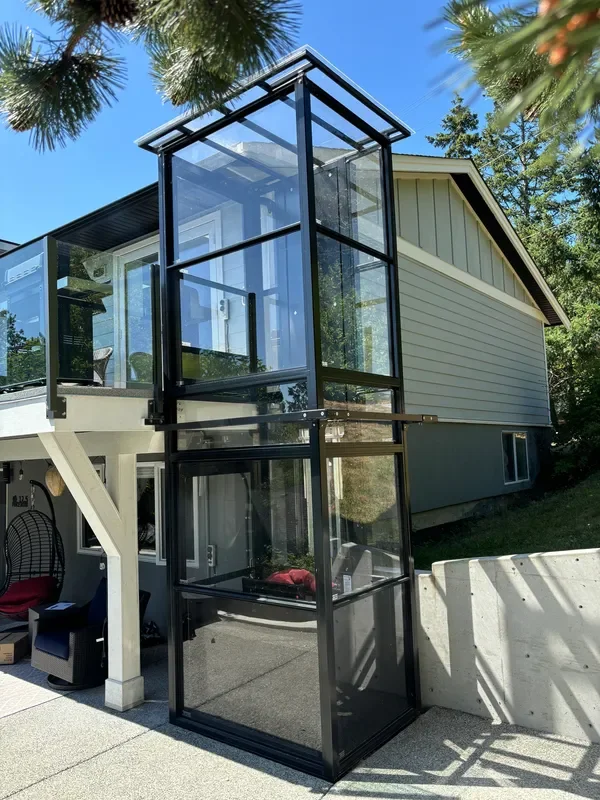 A black-framed glass elevator extending from the ground level to the top of a house's second floor outdoors, with trees and a blue sky in the background.