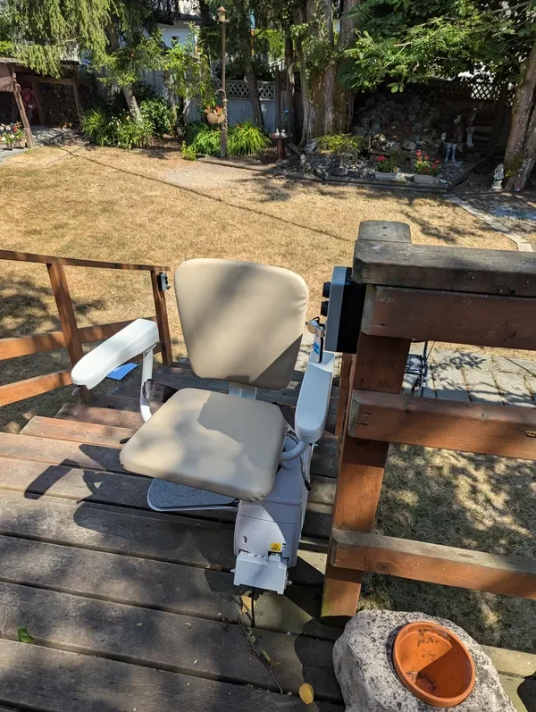 An outdoor wooden deck with a beige lift chair, part of a porch or balcony, overlooking a yard with brown grass, trees, and a garden with flowers.