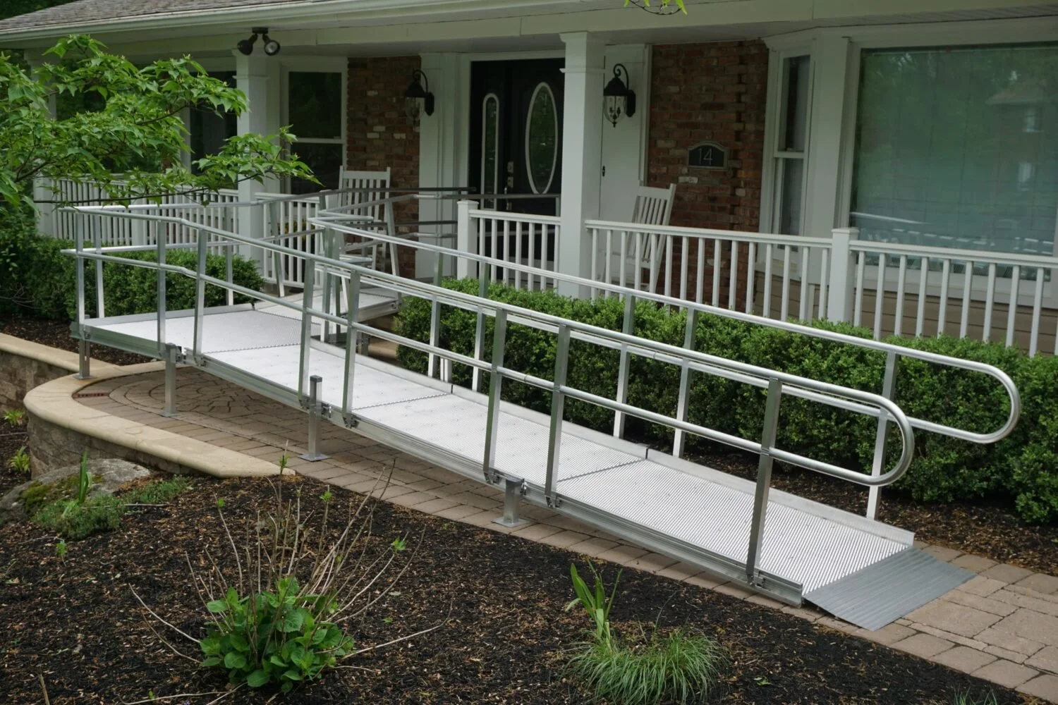 A metal wheelchair ramp leading to a front porch with a white railing, in front of a brick and siding house with a screen door and large windows.