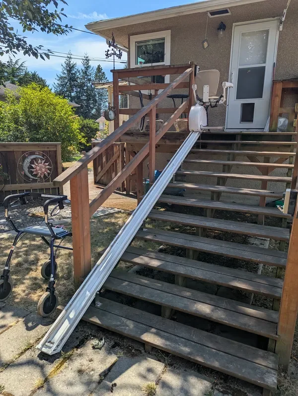 A stair lift with a white seat installed on a wooden staircase outside a house. The stair lift track runs along the steps, and the house has a door and window visible. There is a walker near the stairs and some yard decorations in the background.