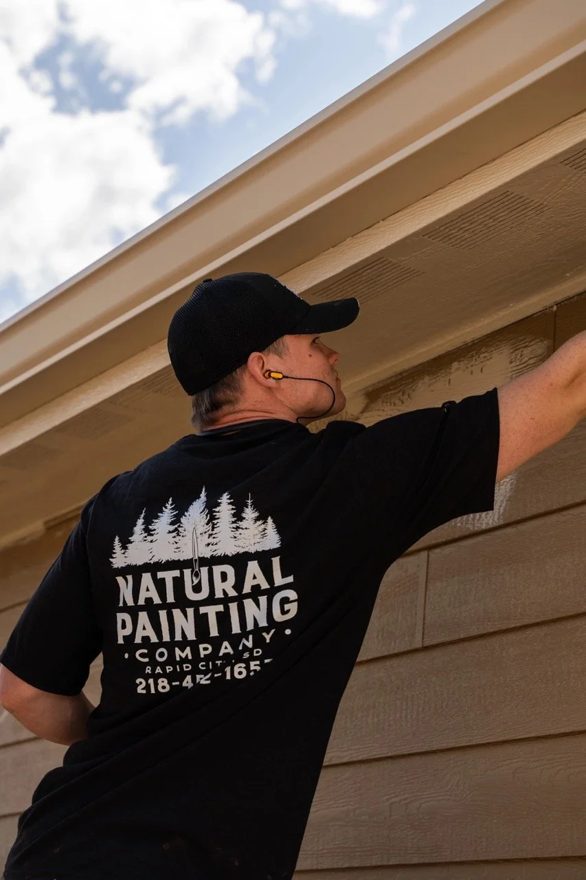 A man in a black cap and black T-shirt with a logo for Natural Painting Company is painting the exterior of a house. The sky is blue with some clouds.