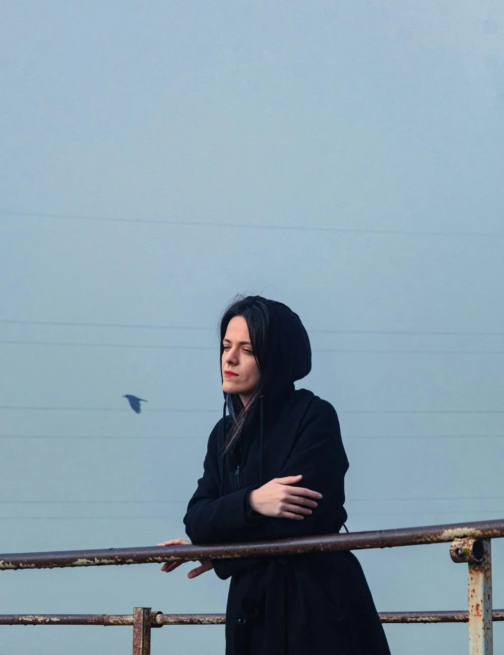 A woman with long dark hair wearing a black hoodie stands near a rusty metal railing outdoors, with her arms crossed and eyes closed, against a hazy sky background.