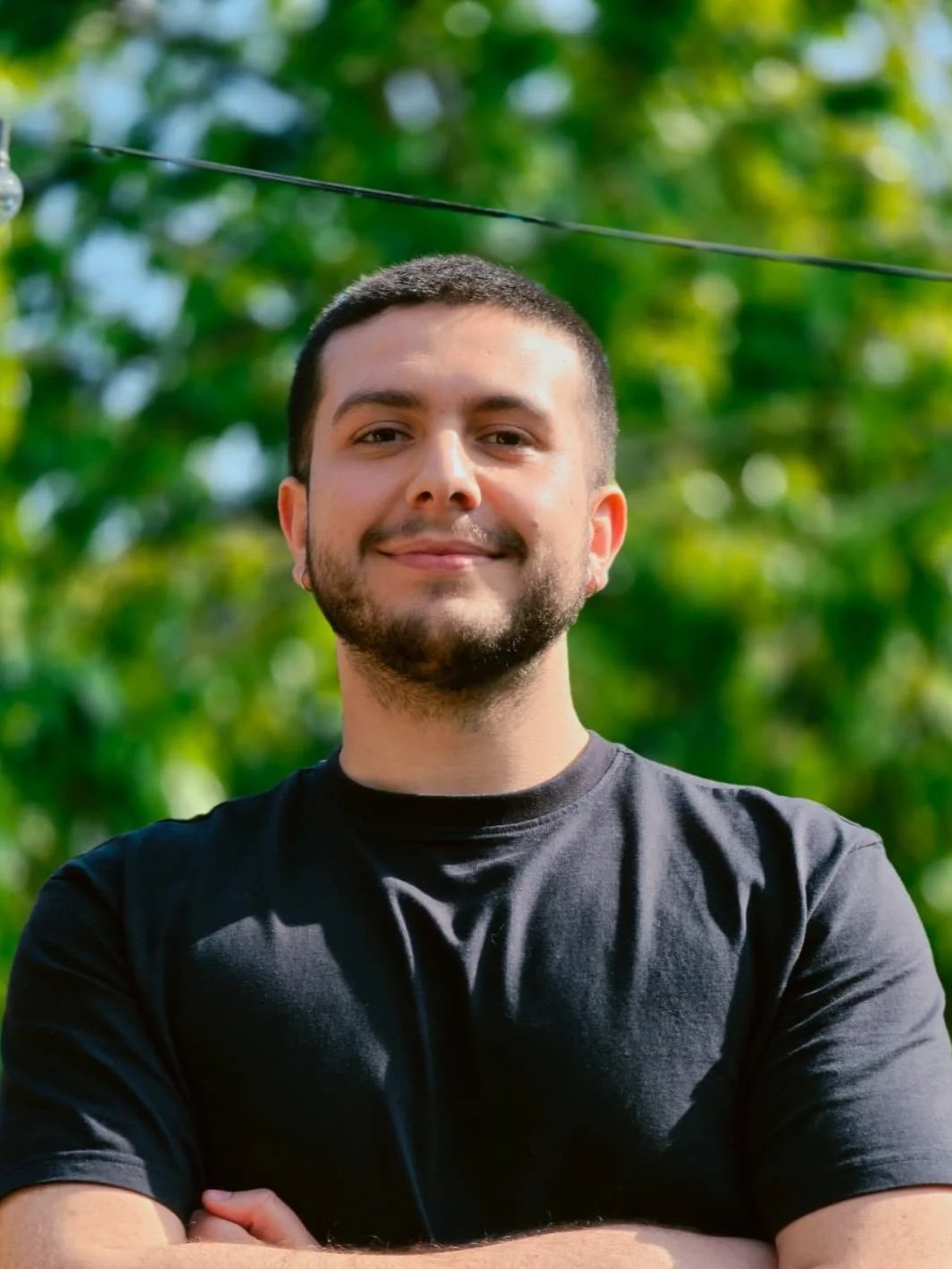 A man with short dark hair and a beard, smiling with arms crossed, standing outdoors in front of green foliage.