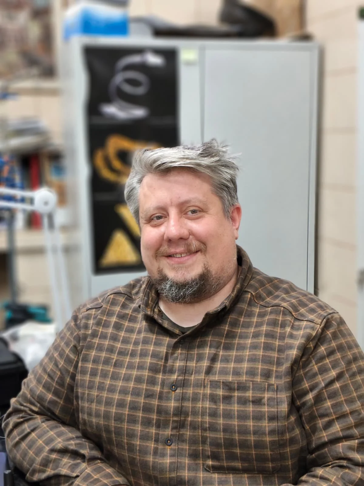 A man with gray hair and a beard smiling at the camera, wearing a brown plaid shirt, seated indoors with a gray cabinet and some shelves with various objects in the background.