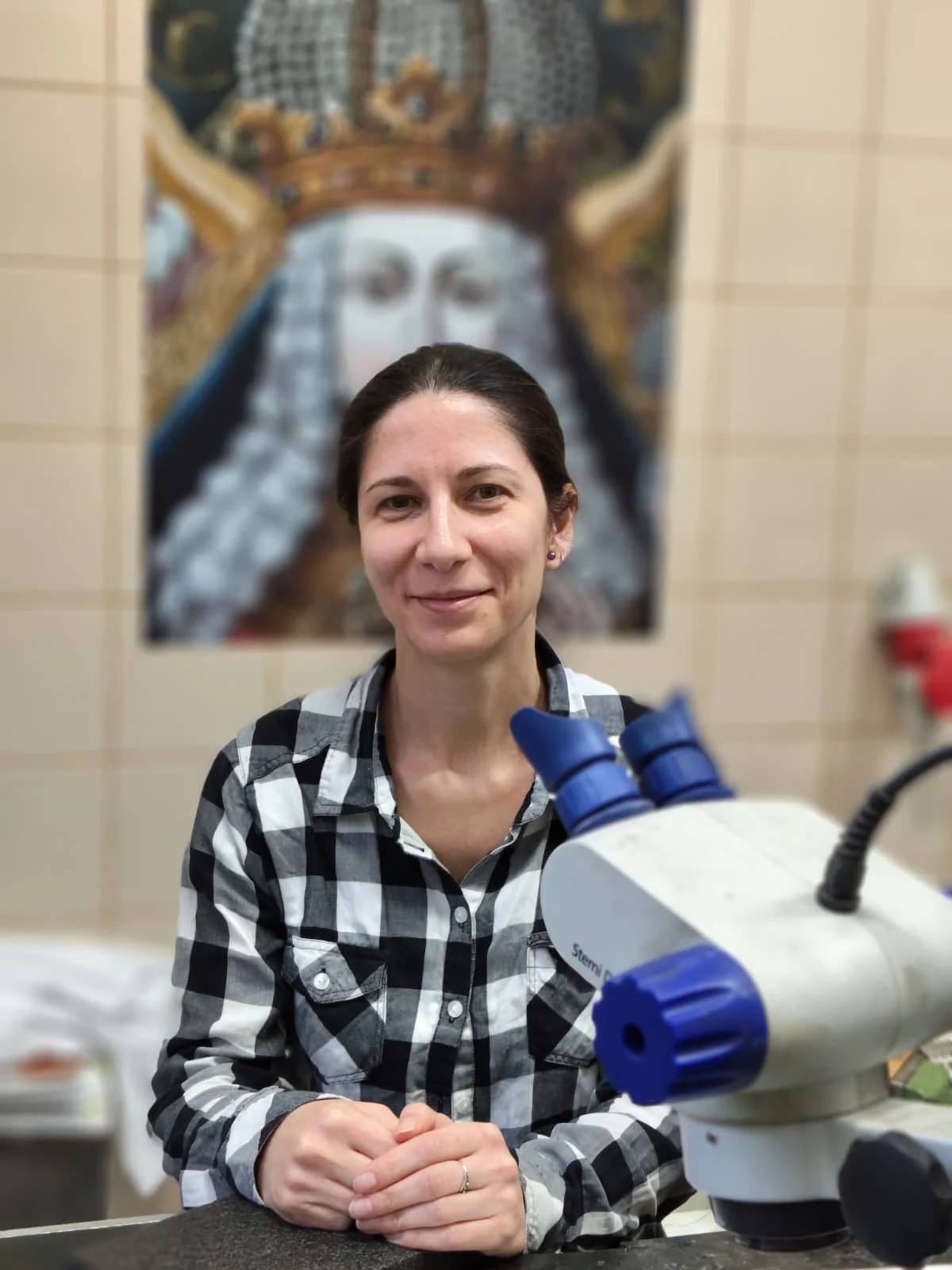 A woman with dark hair and a black-and-white checkered shirt sitting in a laboratory with a microscope in the foreground, a colorful artwork of a regal figure on the wall behind her.