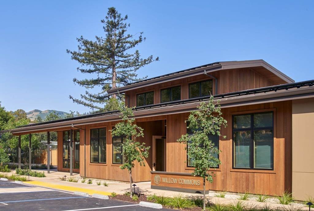 A modern wooden building with large windows, a small sign reading 'WILLOW COMMONS,' surrounded by young trees and landscaped plants, with a parking lot in front and green hills in the background.