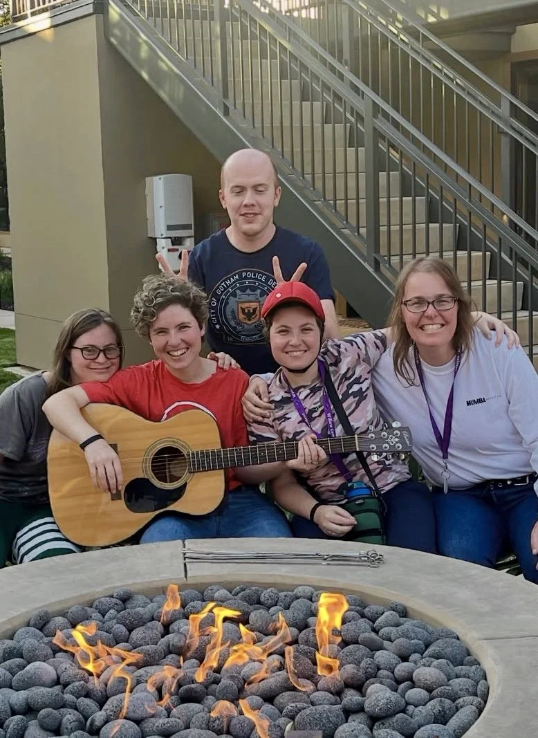 Group of five people sitting outdoors near a fire pit, one person playing guitar, smiling and making peace signs.