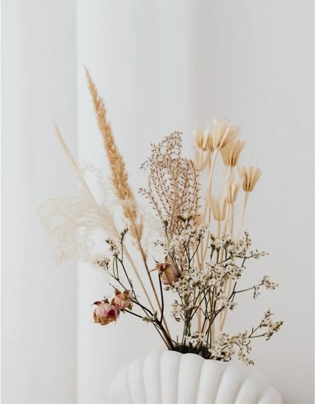 A white vase holding a bouquet of dried flowers, including pampas grass, wheat, and small roses, set against a plain white wall.