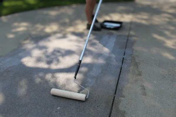 Person using a paint roller to apply gray paint on a concrete surface outdoors.