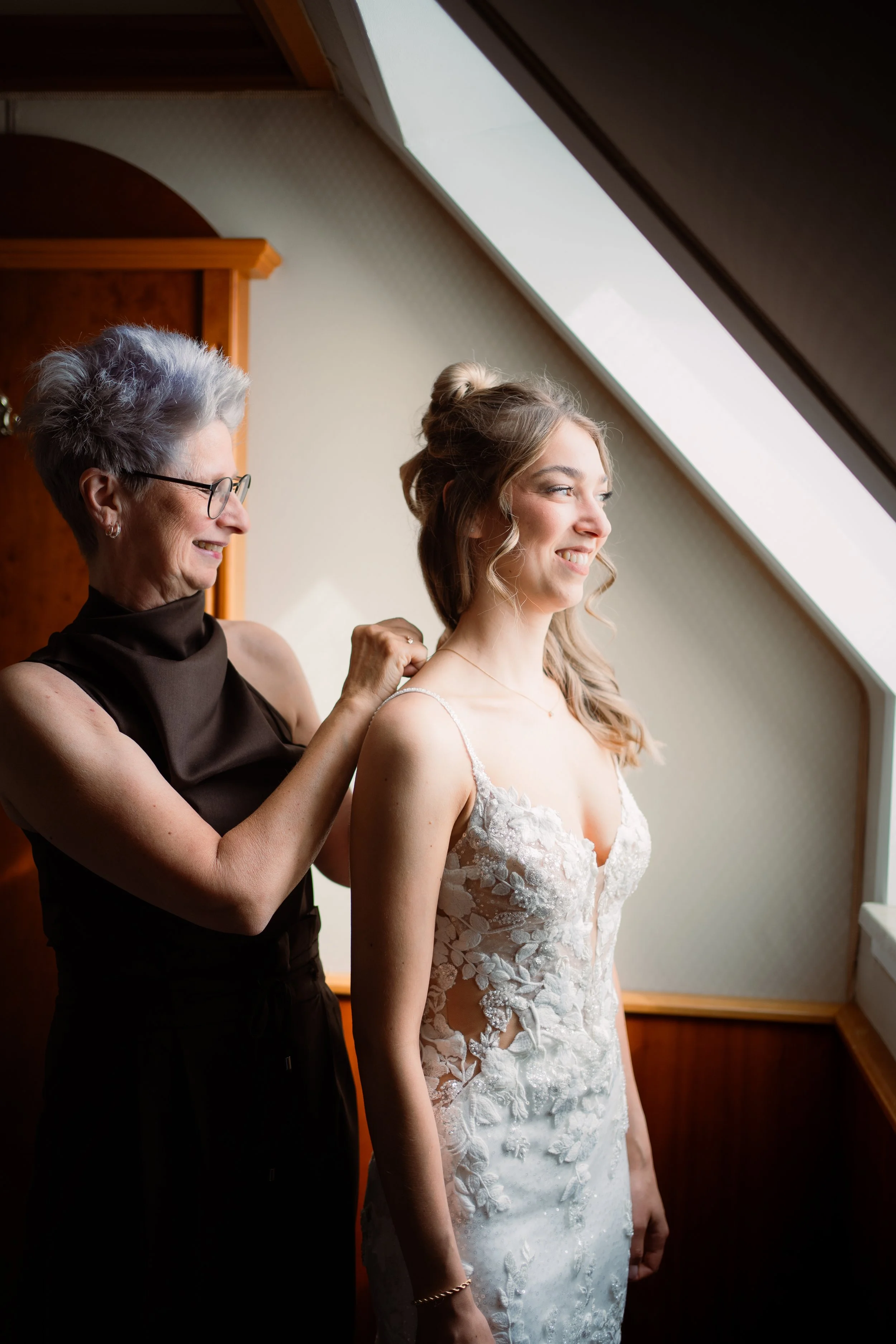 Eine ältere Frau hilft einer jungen Frau, ein Hochzeitskleid anzuprobieren, vor einem großen Dachfenster