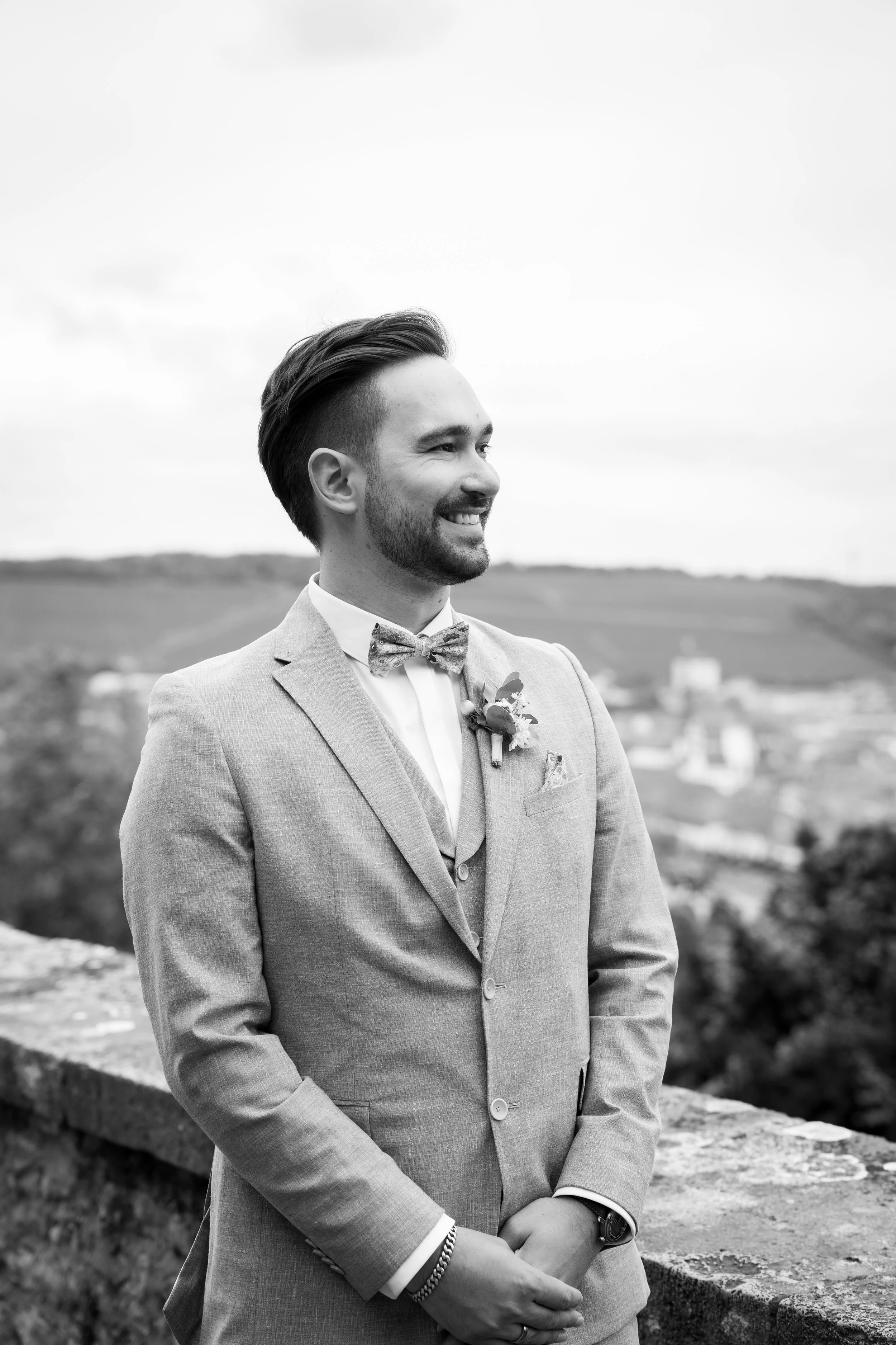 Ein lächelnder Mann in formeller Kleidung und Fliege steht im Freien an einer Mauer, im Hintergrund eine ländliche Landschaft mit Hügeln und Gebäuden, in Schwarz-Weiß aufgenommen.