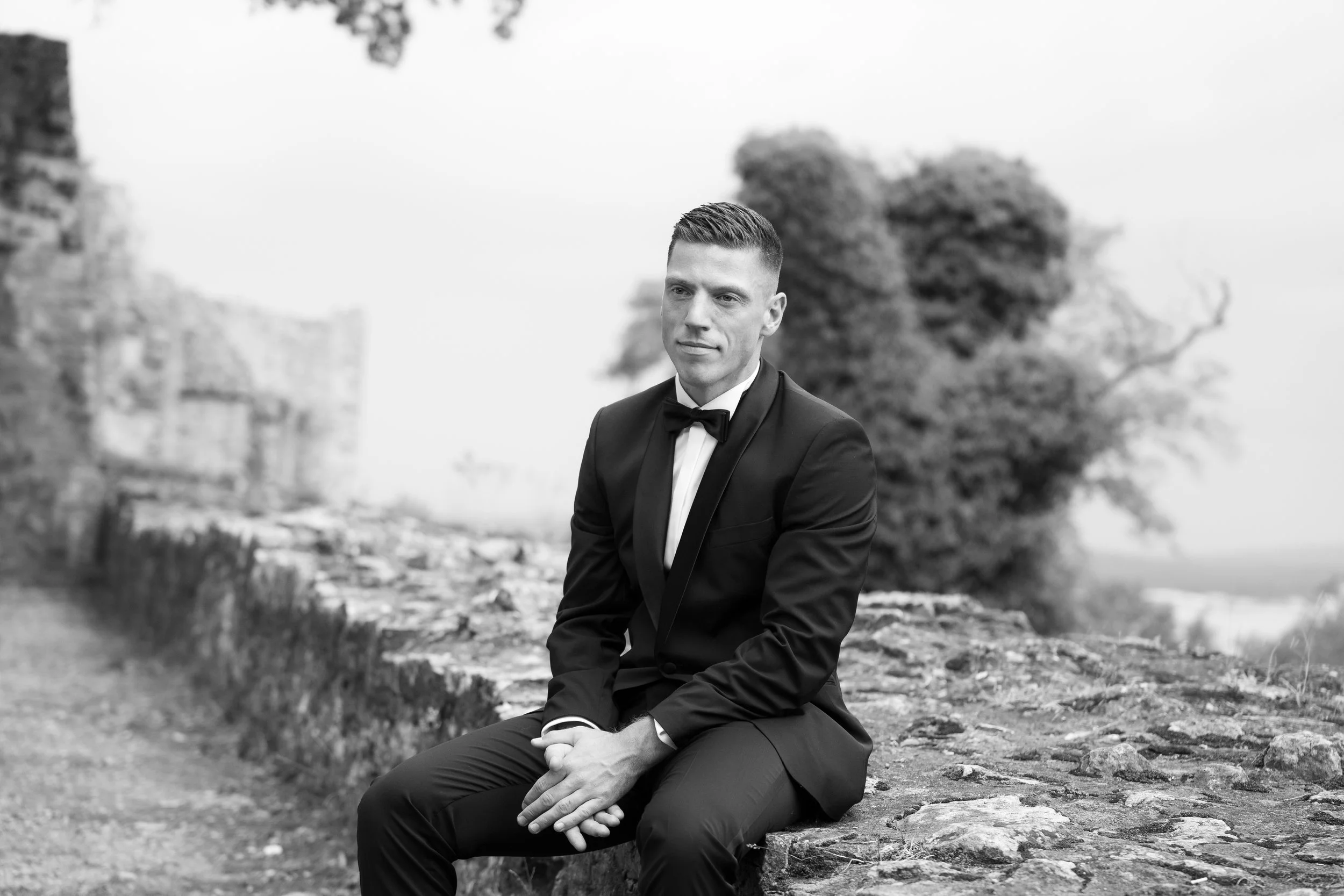 Ein junger Mann in einem eleganten schwarzen Anzug mit Fliege sitzt auf einem Felsen im Freien, im Hintergrund sind Bäume und eine unscharfe Landschaft zu sehen, schwarz-weiß Aufnahme.