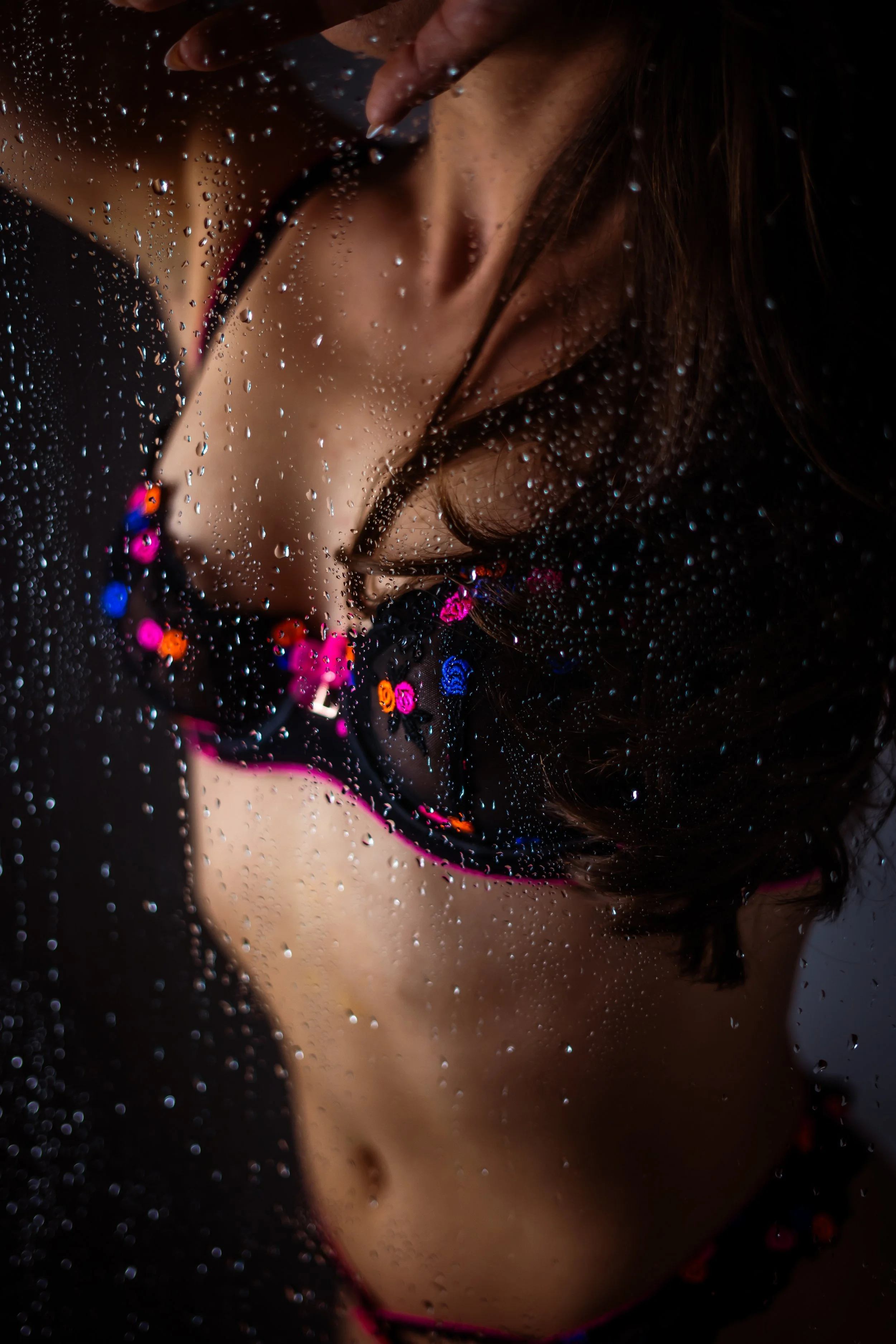 Nahaufnahme eines Mädchens, das durch eine regennasse Glasscheibe betrachtet wird, verziert mit Wassertröpfchen, trägt bunte Halsketten und trägt ein dunkles Oberteil mit pinken Akzenten. Boudoir, Akt, Sinnliche Fotoshooting, Senusalshooting