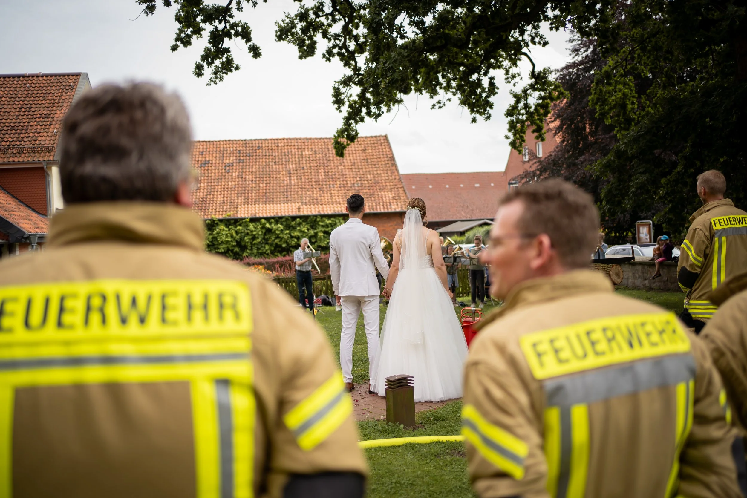 Feuerwehrleute im Vordergrund, bei einem Hochzeitszeremonie im Freien im Hintergrund, mit Braut und Bräutigam, an einem Tag mit bewölktem Himmel, umgeben von Bäumen und alten Gebäuden.