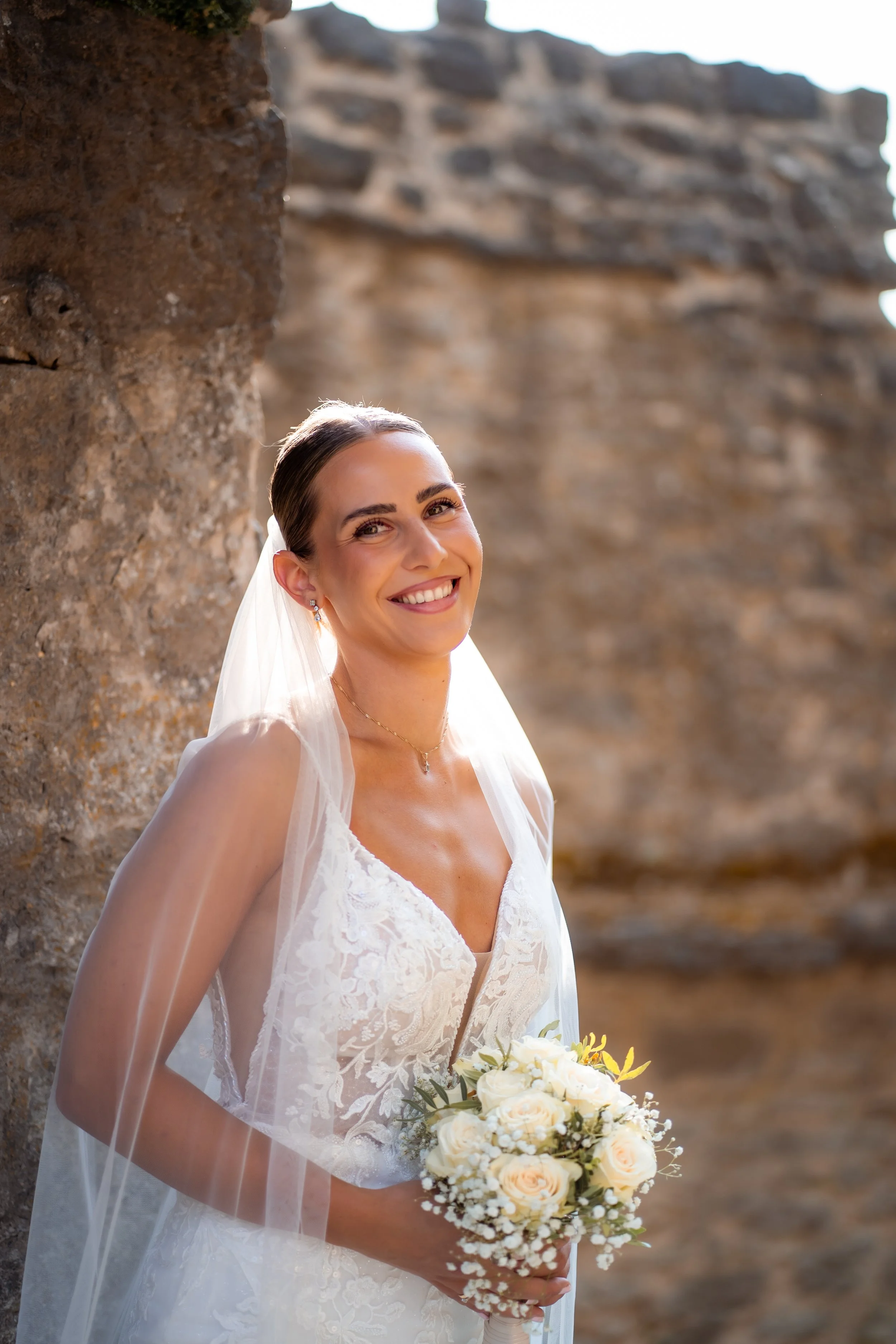 Eine lächelnde Braut in einem weißen Brautkleid mit Blumenstrauß vor einer alten Steinmauer.