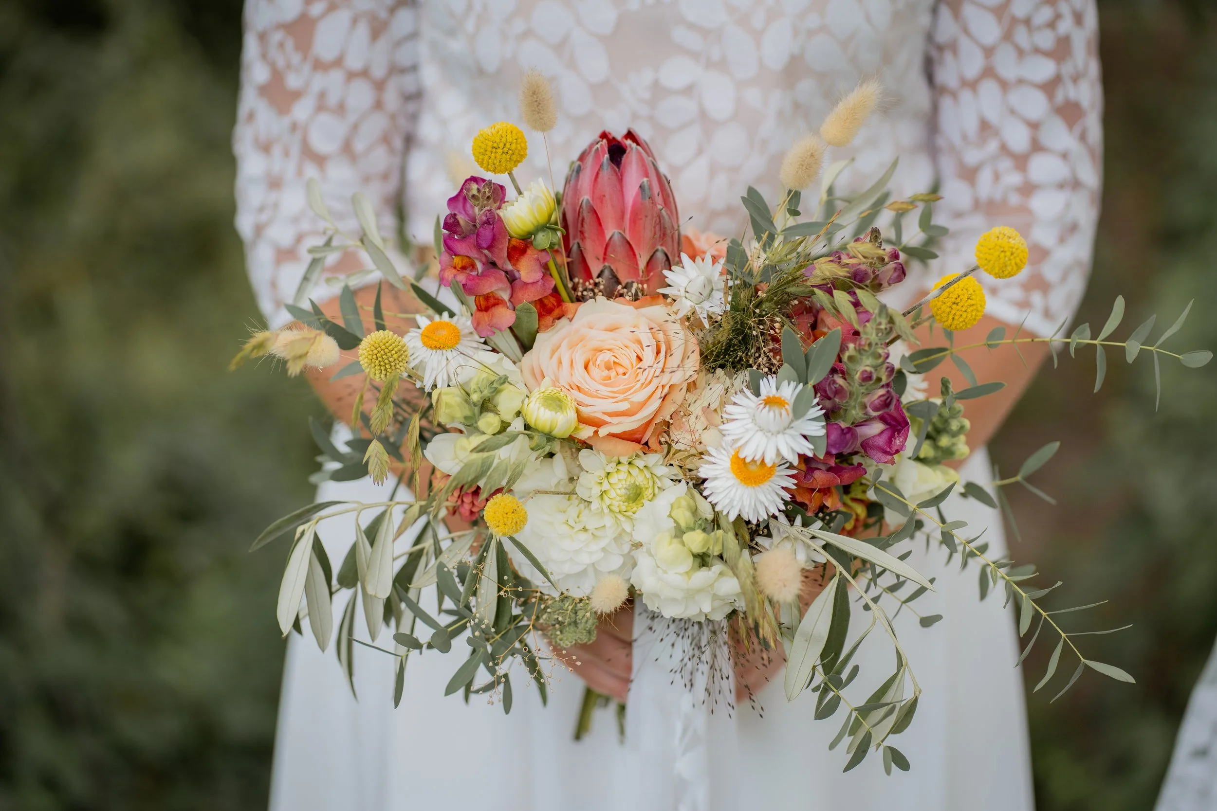 Hochzeitsstrauß mit roten, weißen, gelben und lila Blumen, verbunden mit grünen Blättern und silbernen Zweigen, vor einer Person in einem weißen Kleid mit Spitzenmuster.