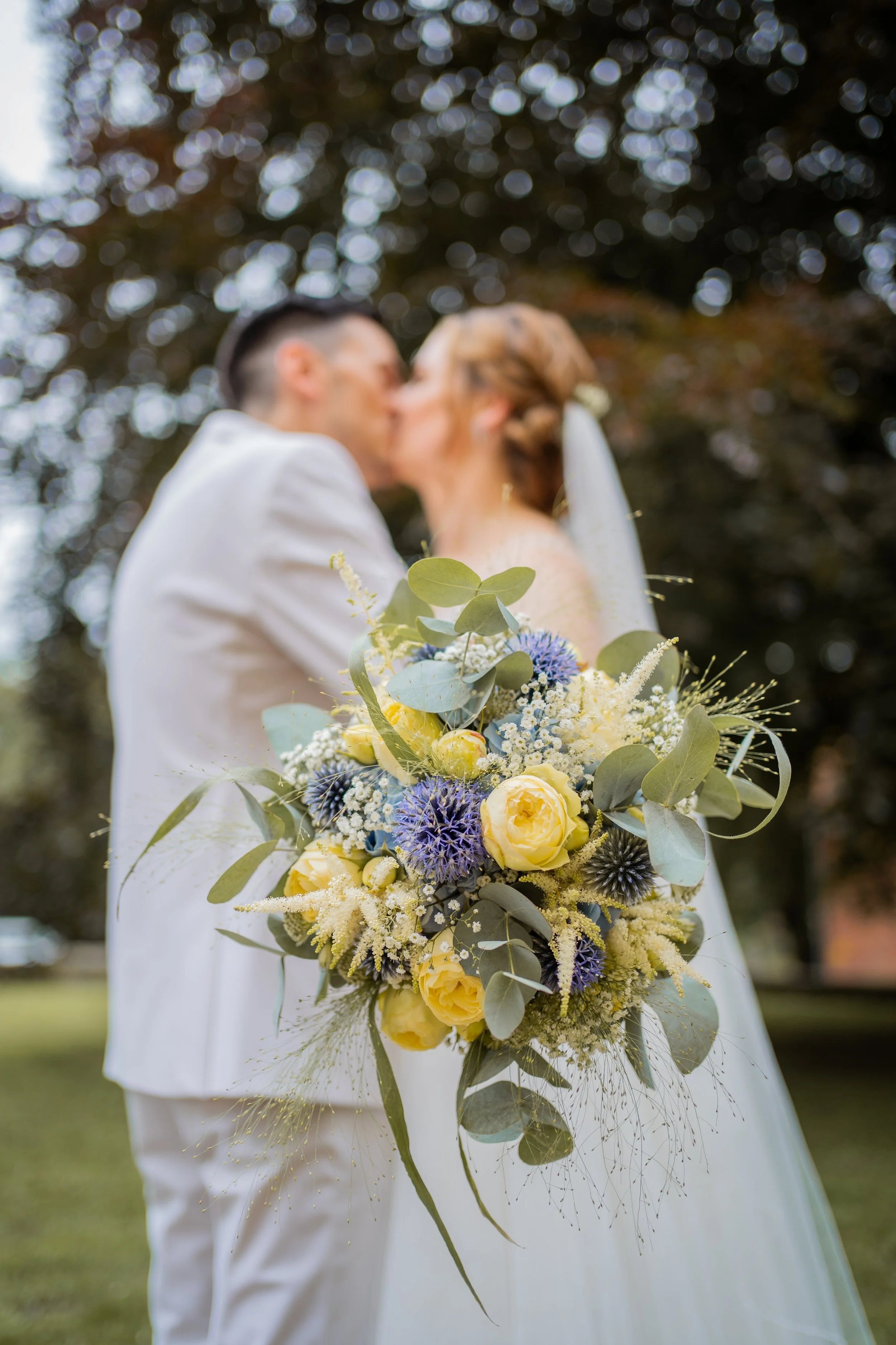 Ein Brautpaar küsst sich bei einer Hochzeit im Freien, mit Blumenstrauß im Vordergrund. Die Szene ist unscharf im Hintergrund mit Bäumen und Sonnenlicht.