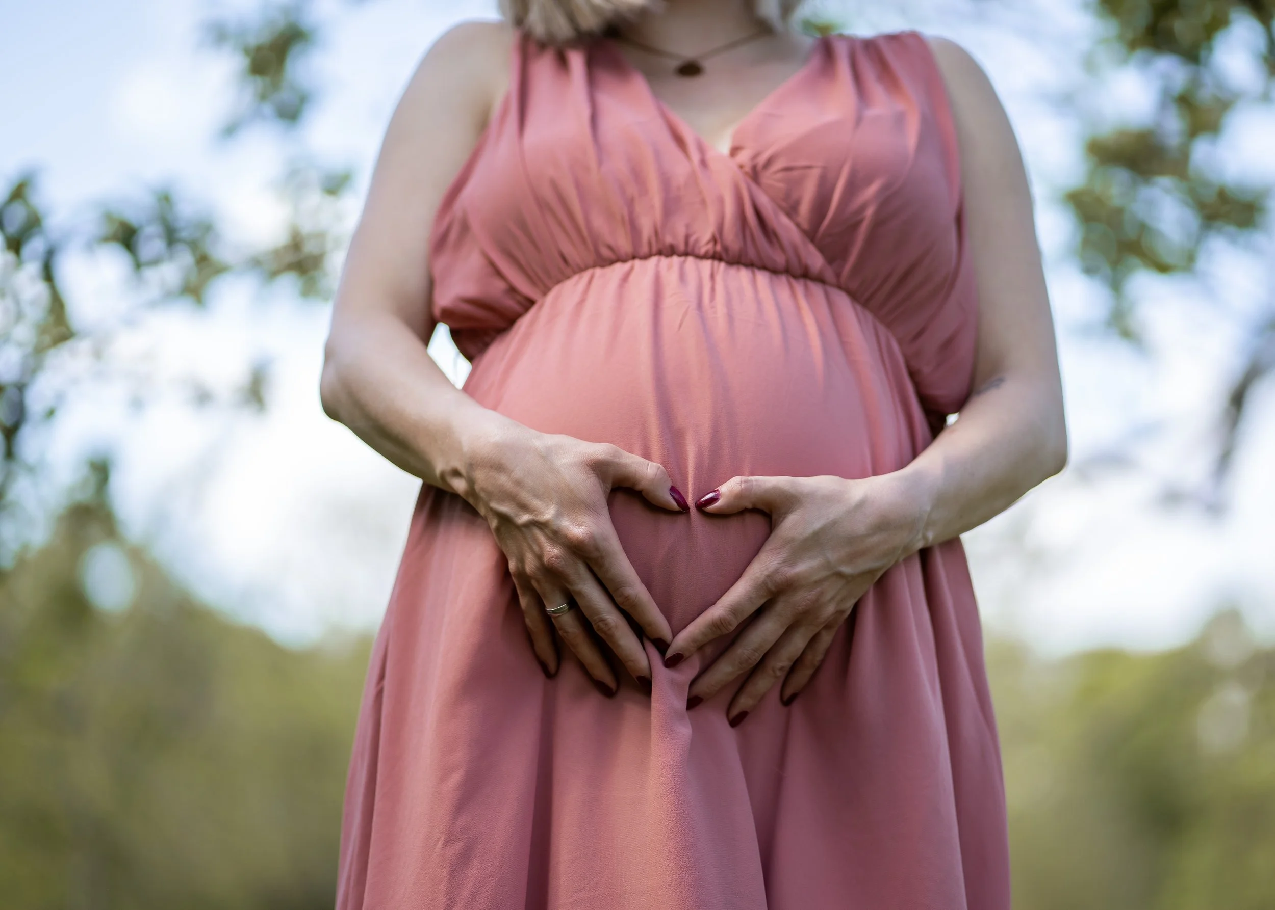 Eine schwangere Frau in einem pinkfarbenen Kleid macht eine Herzform mit ihren Händen auf ihrem Bauch, im Hintergrund sind Bäume und Himmel zu sehen.