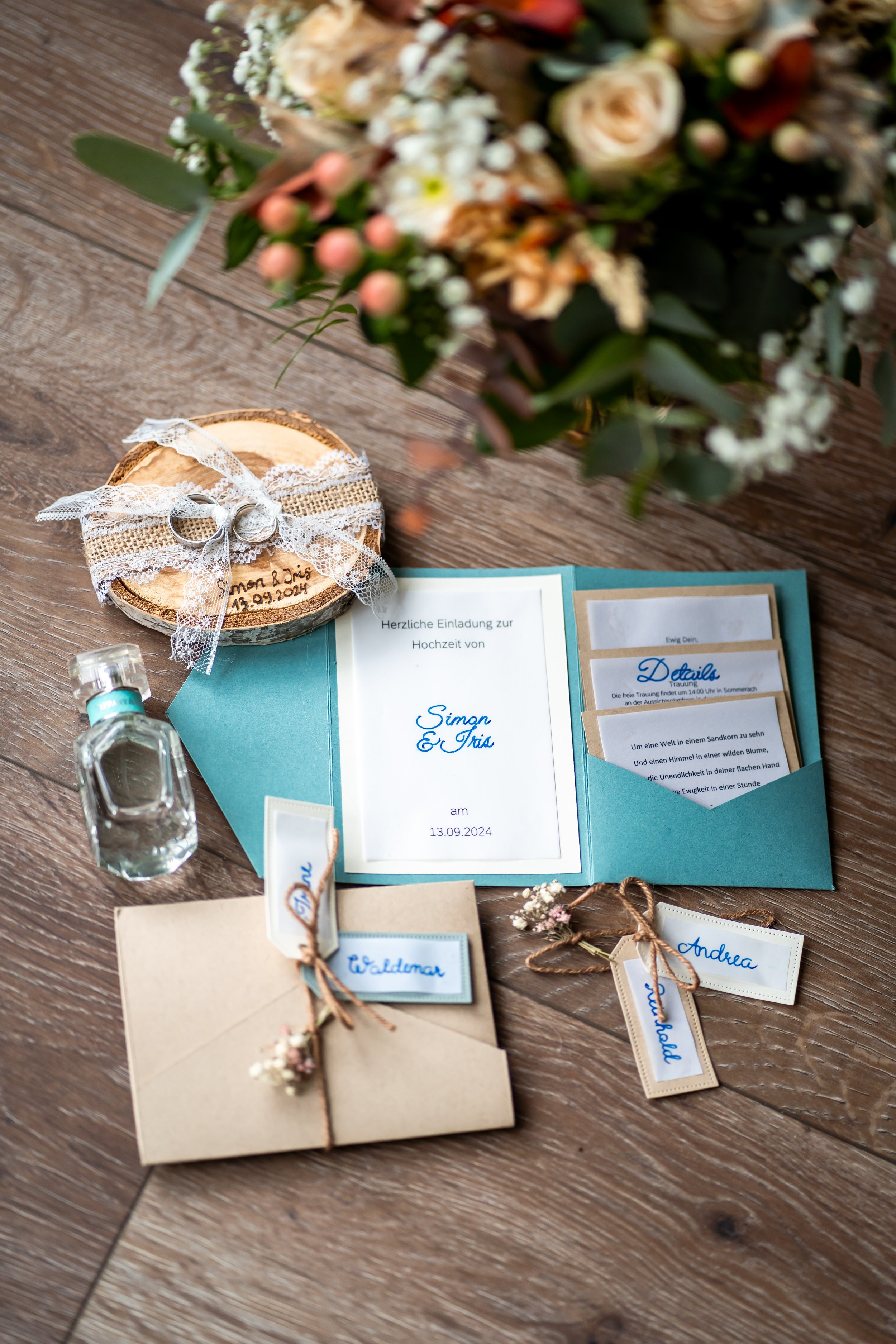Blumenstrauß auf einem Tisch mit Hochzeits- oder Einladungskarten, kleinen Geschenken und Wasserflaschen, nahe bei einer Holzscheibe mit eingefrästem Datum und Namen.