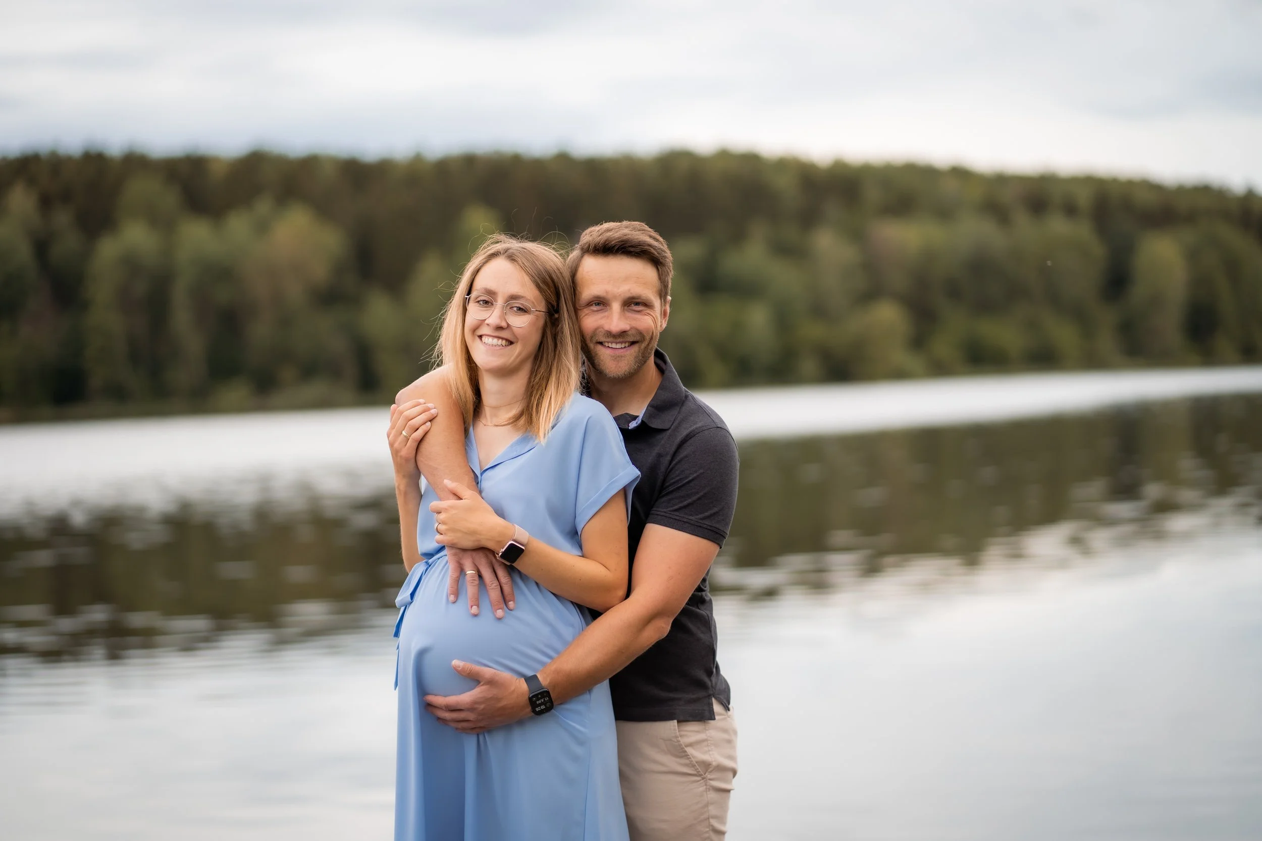 Ein glückliches Paar am Fluss, die schwangere Frau trägt ein blaues Kleid, der Mann umarmt sie aus hinter ihr, um sie zu unterstützen, im Hintergrund sind Bäume und bewölkter Himmel.