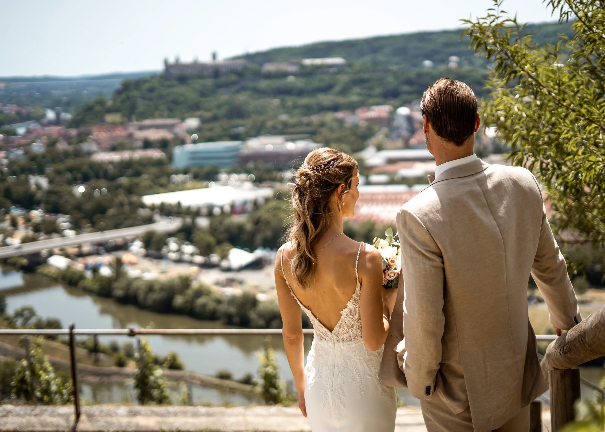 Ein Brautpaar, ein Mann und eine Frau, steht draußen mit Blick auf eine hügelige Landschaft mit einem Fluss. Der Mann trägt einen hellen Anzug, die Frau ein weißes Hochzeitskleid.